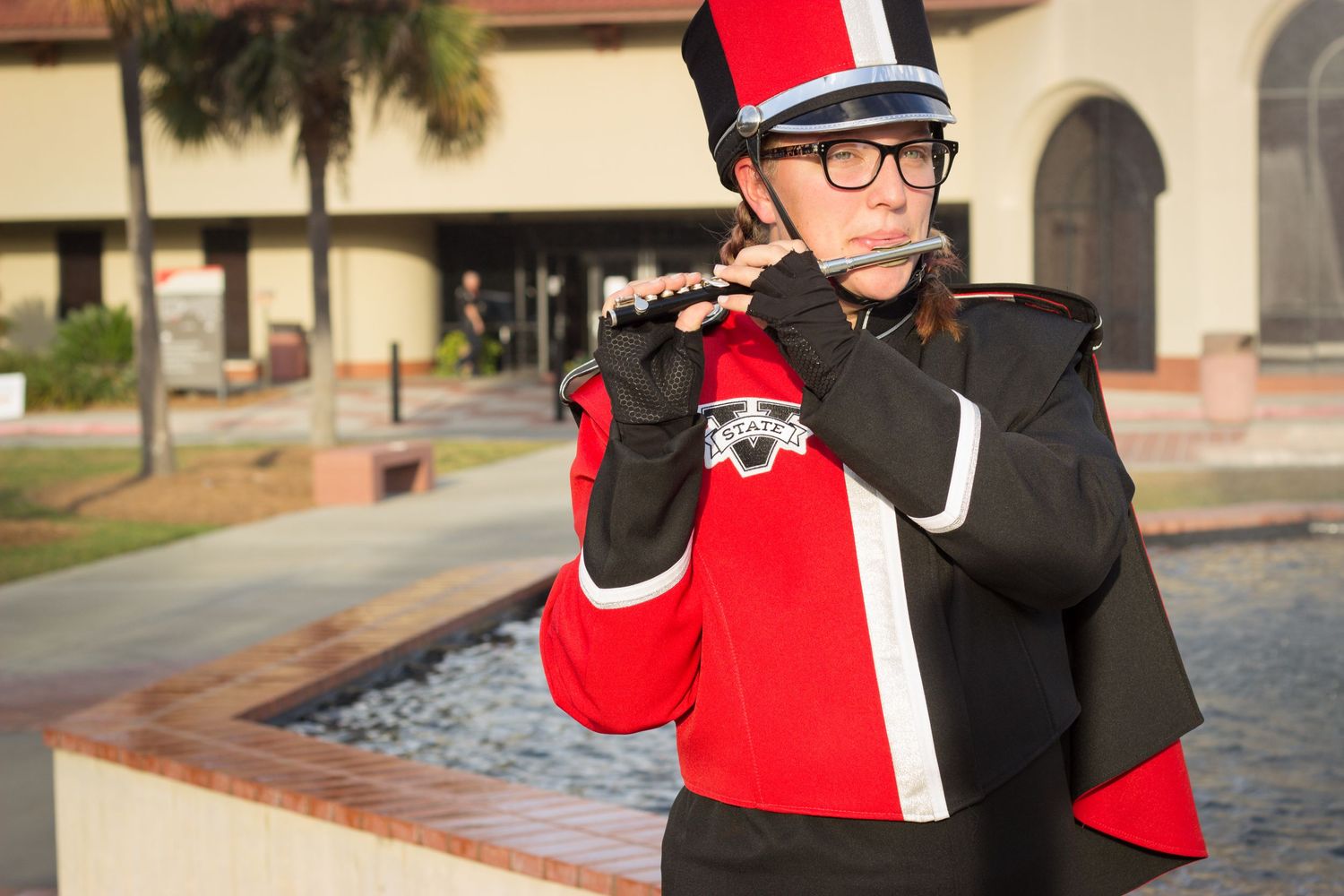 Marching band member in red and black uniform playing flute outdoors near fountain.