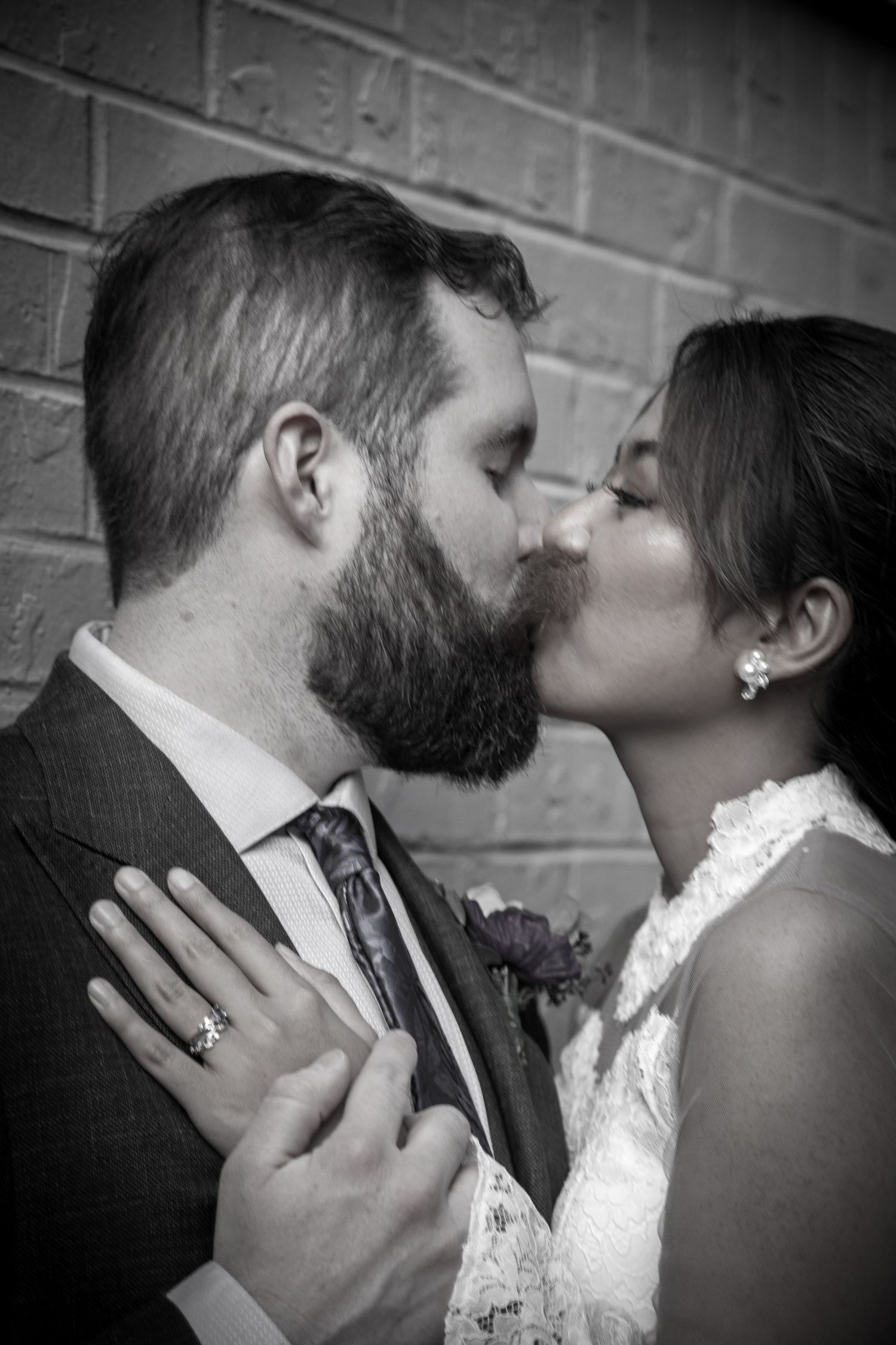 Black and white close-up of bride and bearded groom sharing a kiss against brick wall.