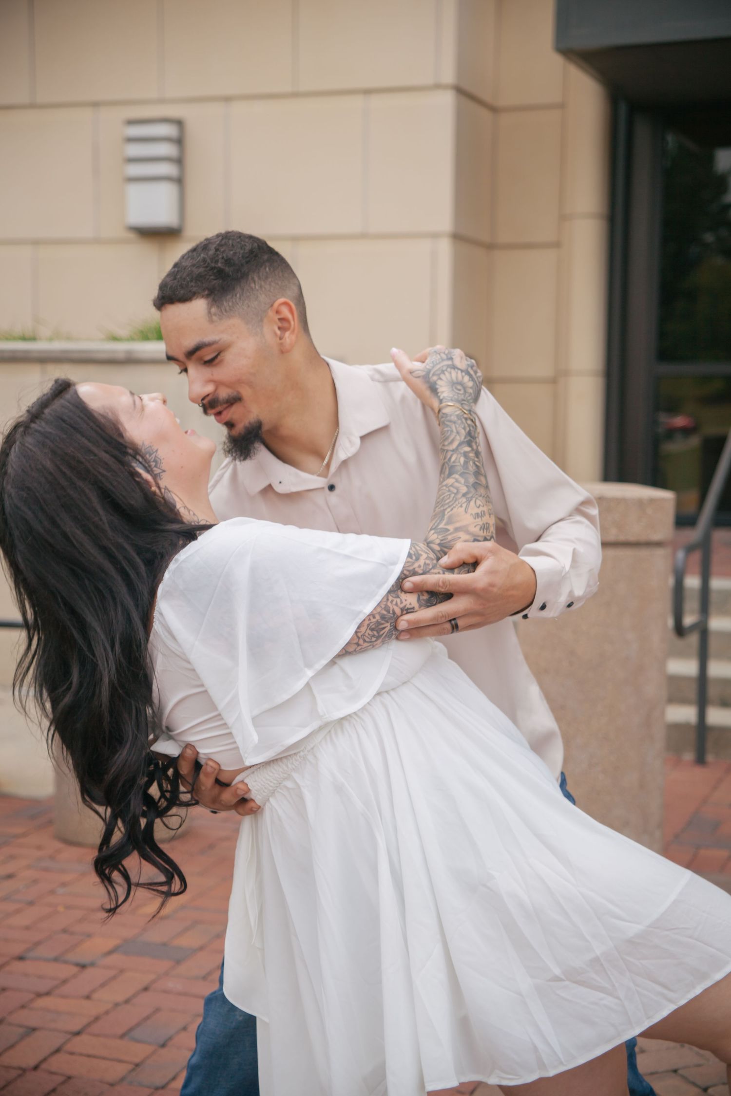 Couple dancing outdoors, tattooed man dipping woman in white dress on brick patio.