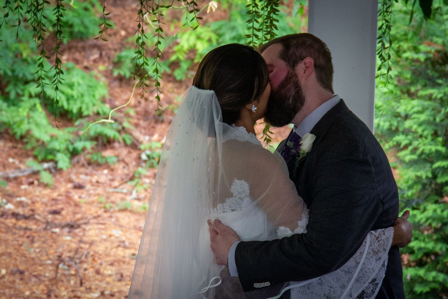 Newlywed couple sharing first kiss under weeping willow branches at outdoor garden wedding ceremony.