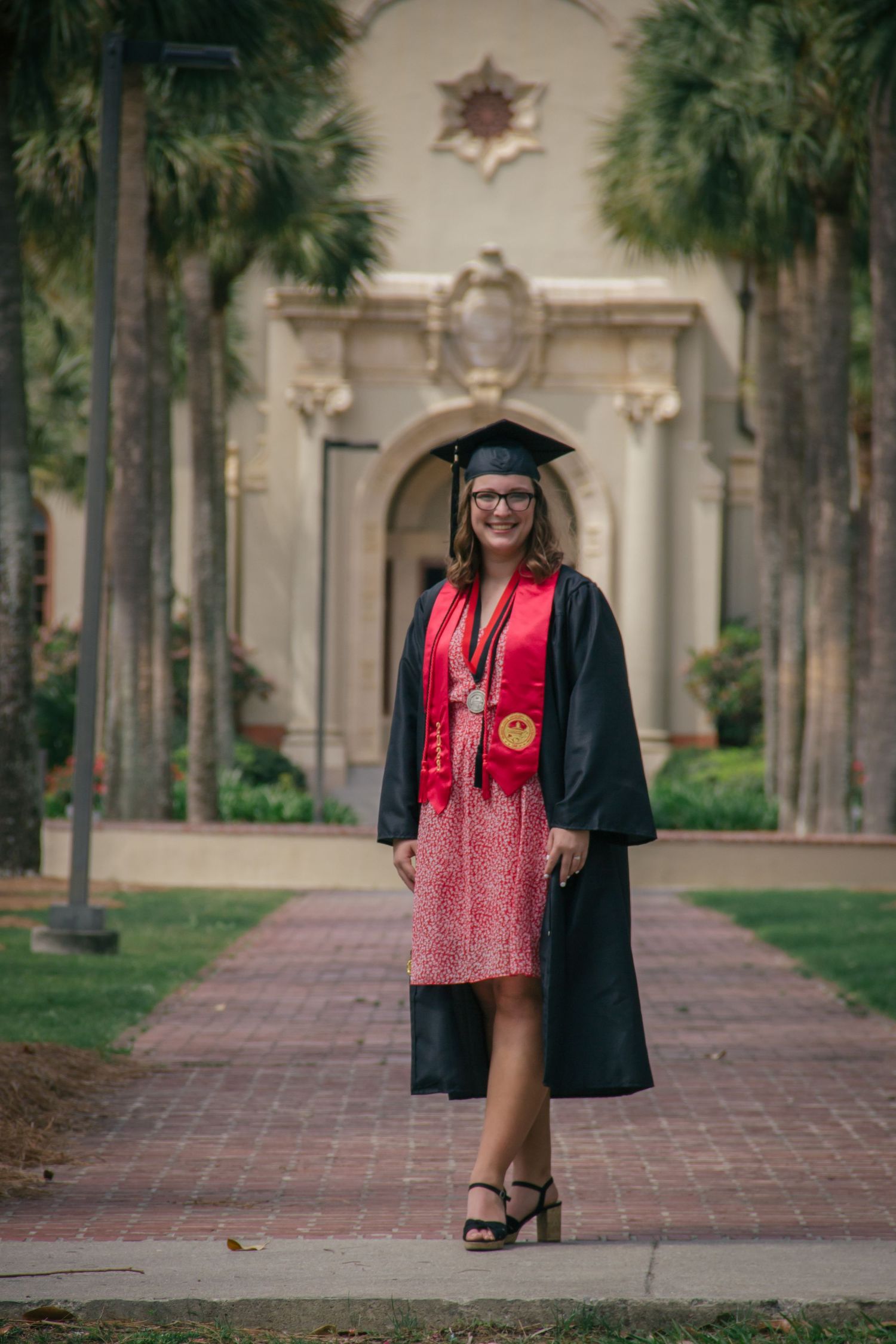 Female graduate in cap and gown with red stole posing on brick path lined with palm trees.