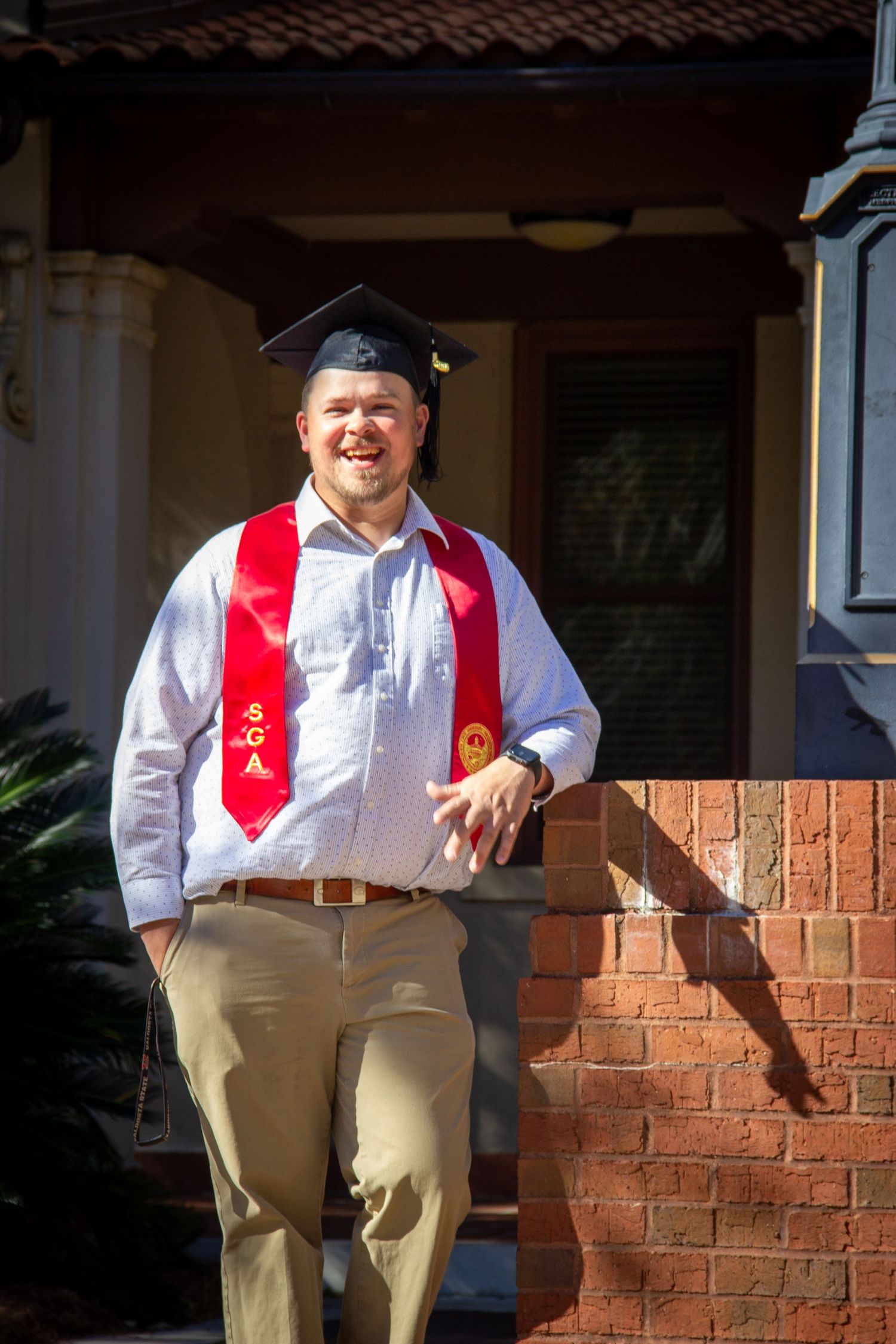Laughing graduate in cap and red stole leaning on brick wall outside campus building.
