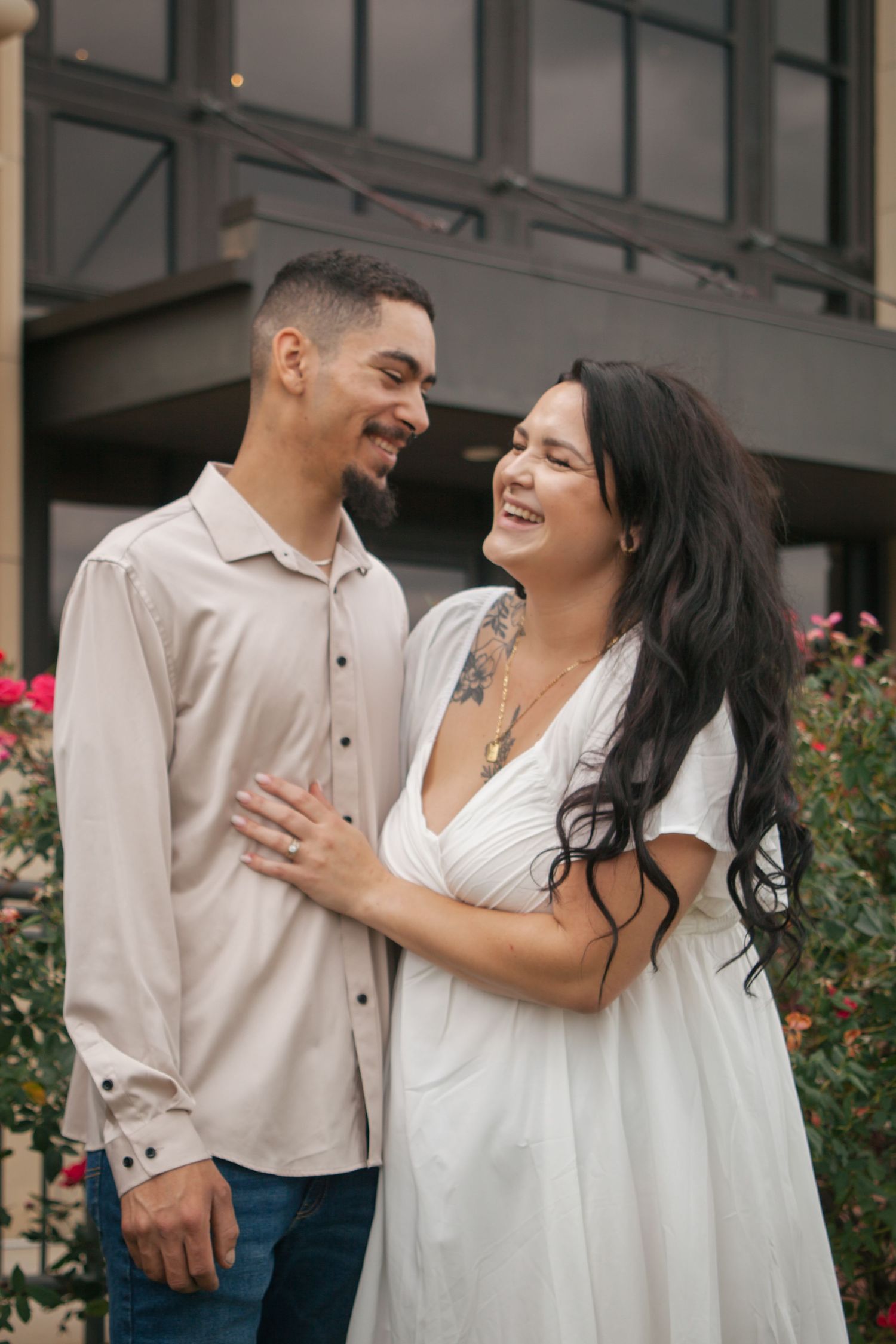 Laughing tattooed woman in white dress embraces smiling man in beige shirt during outdoor engagement session.