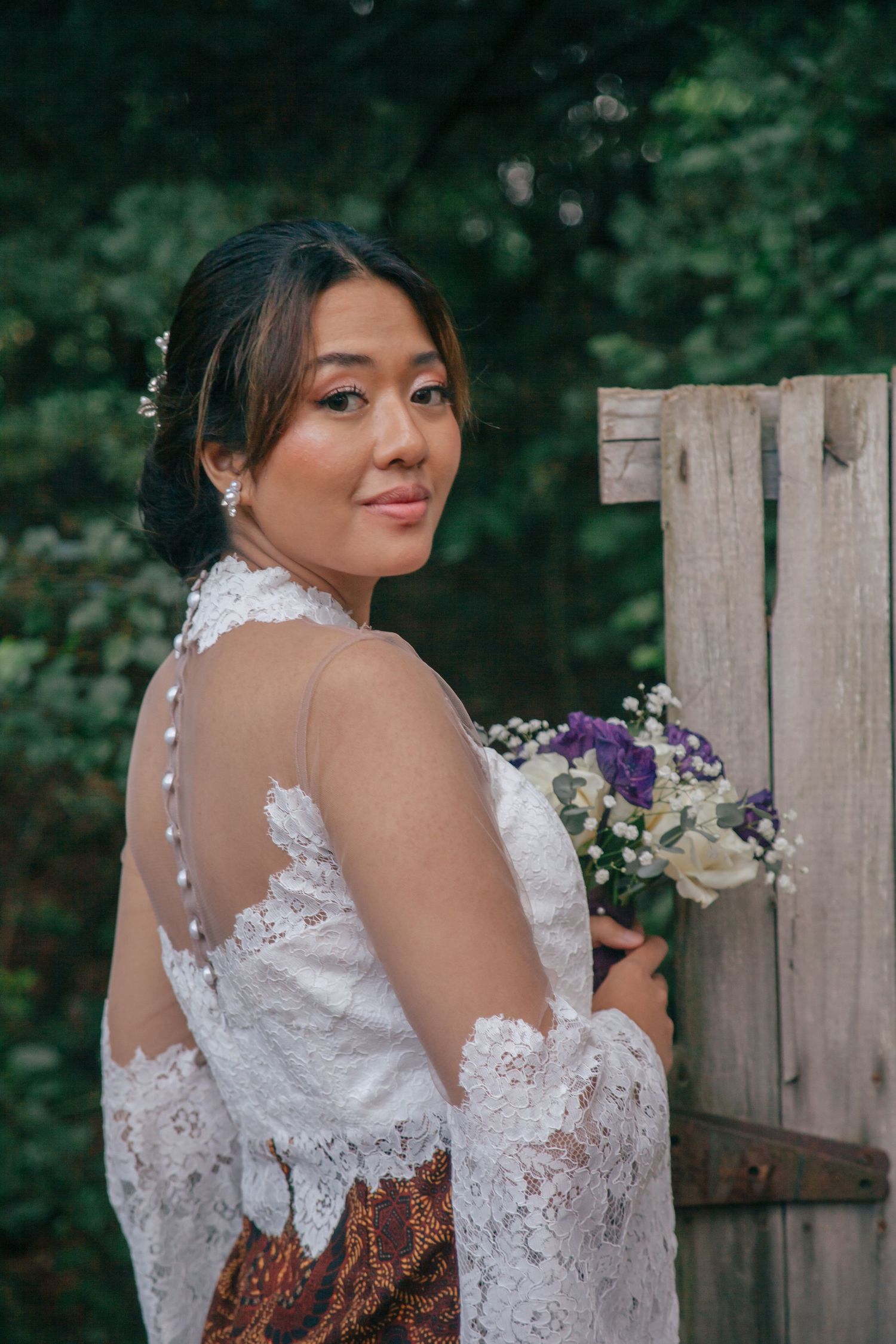 Elegant bride in lace off-shoulder gown holding purple and white bouquet beside a rustic wooden fence.
