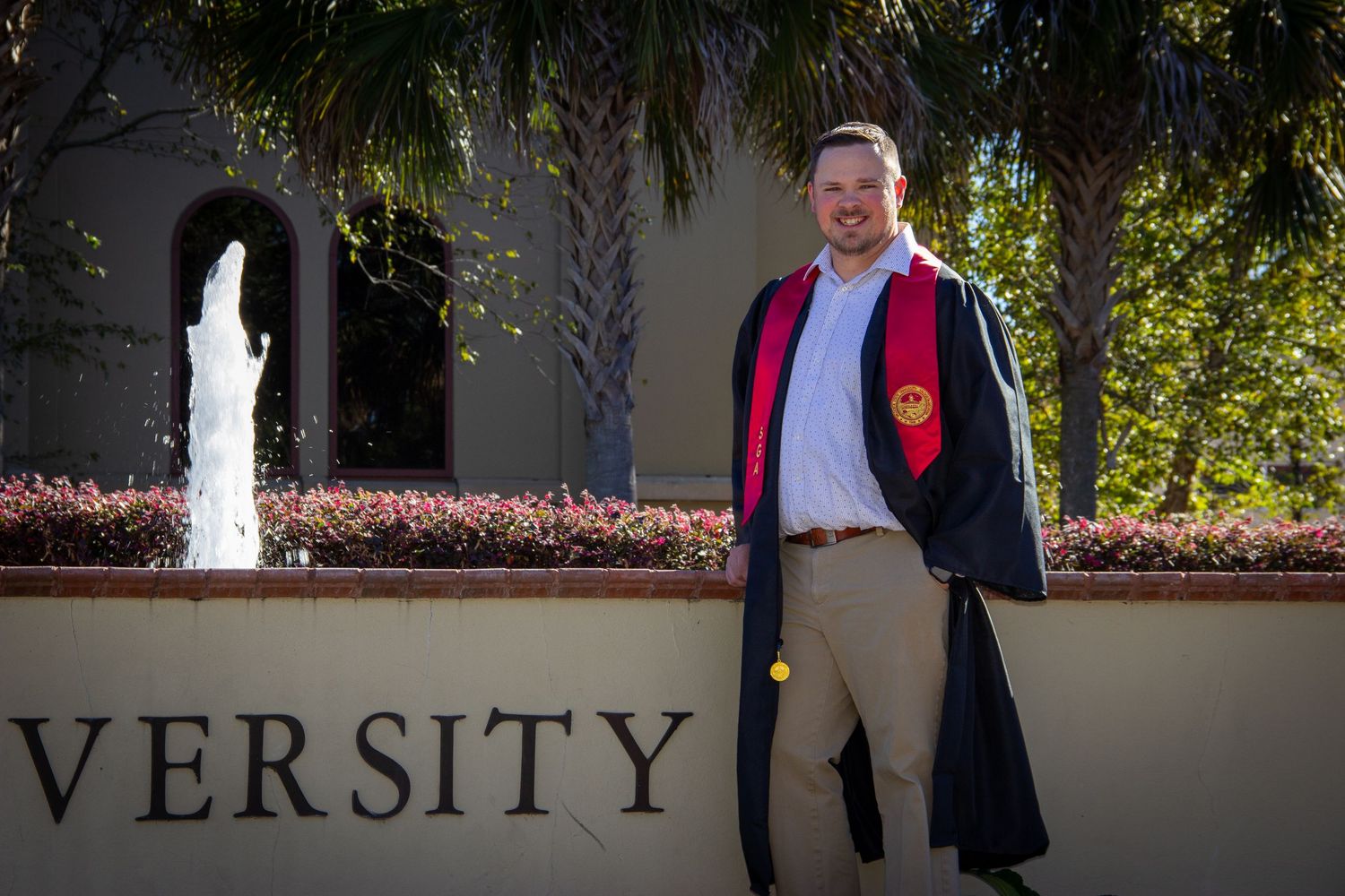 Smiling graduate in cap and gown posing beside university sign with fountain backdrop.