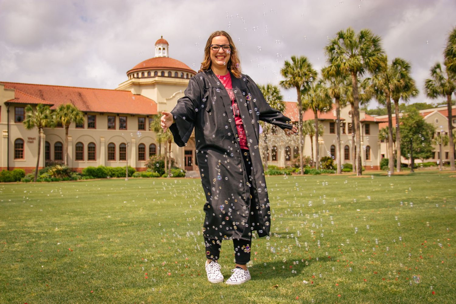 Graduate in black gown celebrating on green lawn in front of historic university building.