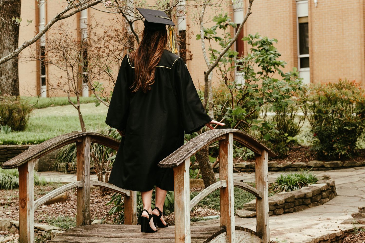 Female graduate in black cap and gown walking across wooden bridge on college campus.
