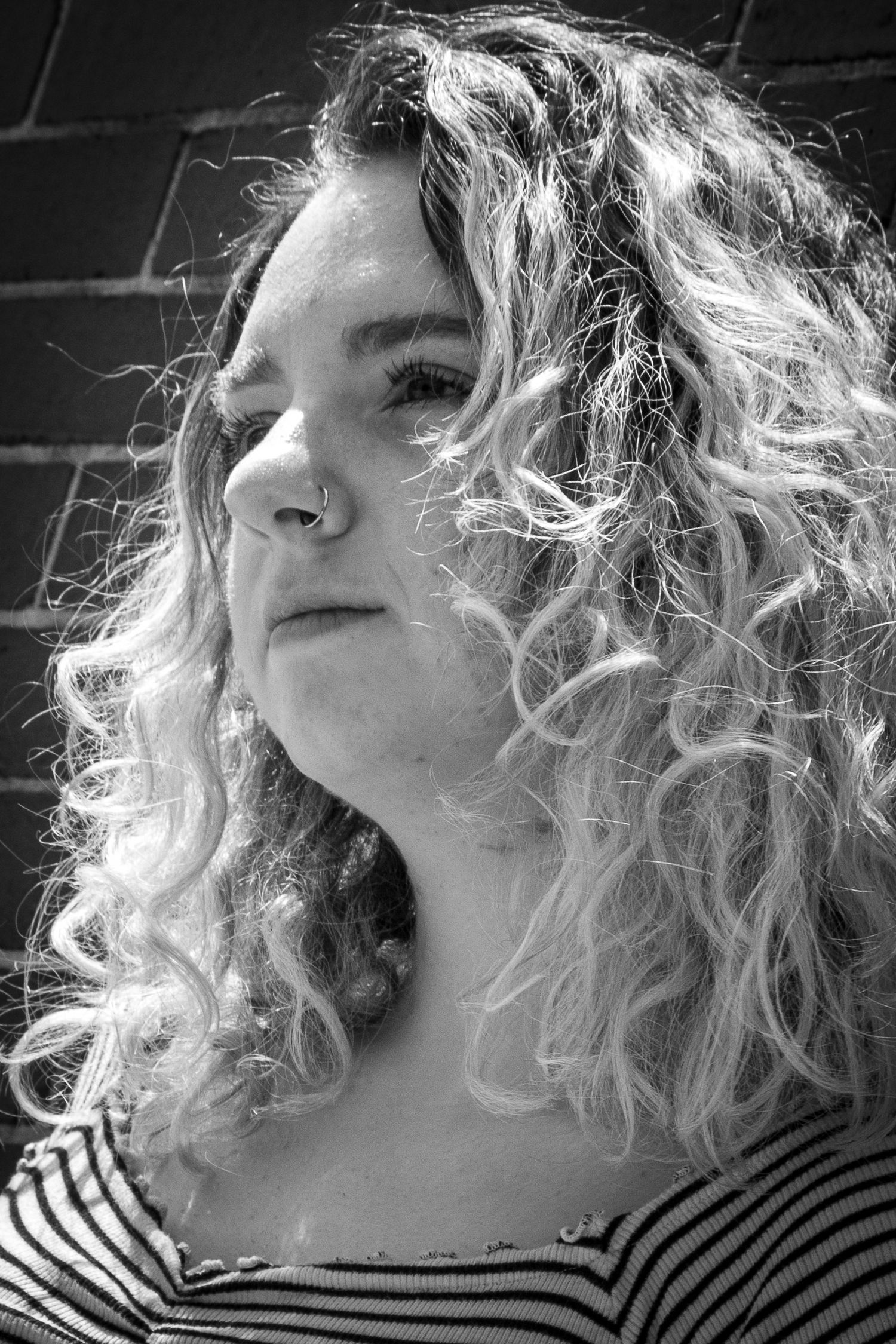 Black and white close-up portrait of young woman with curly blonde hair against brick wall.
