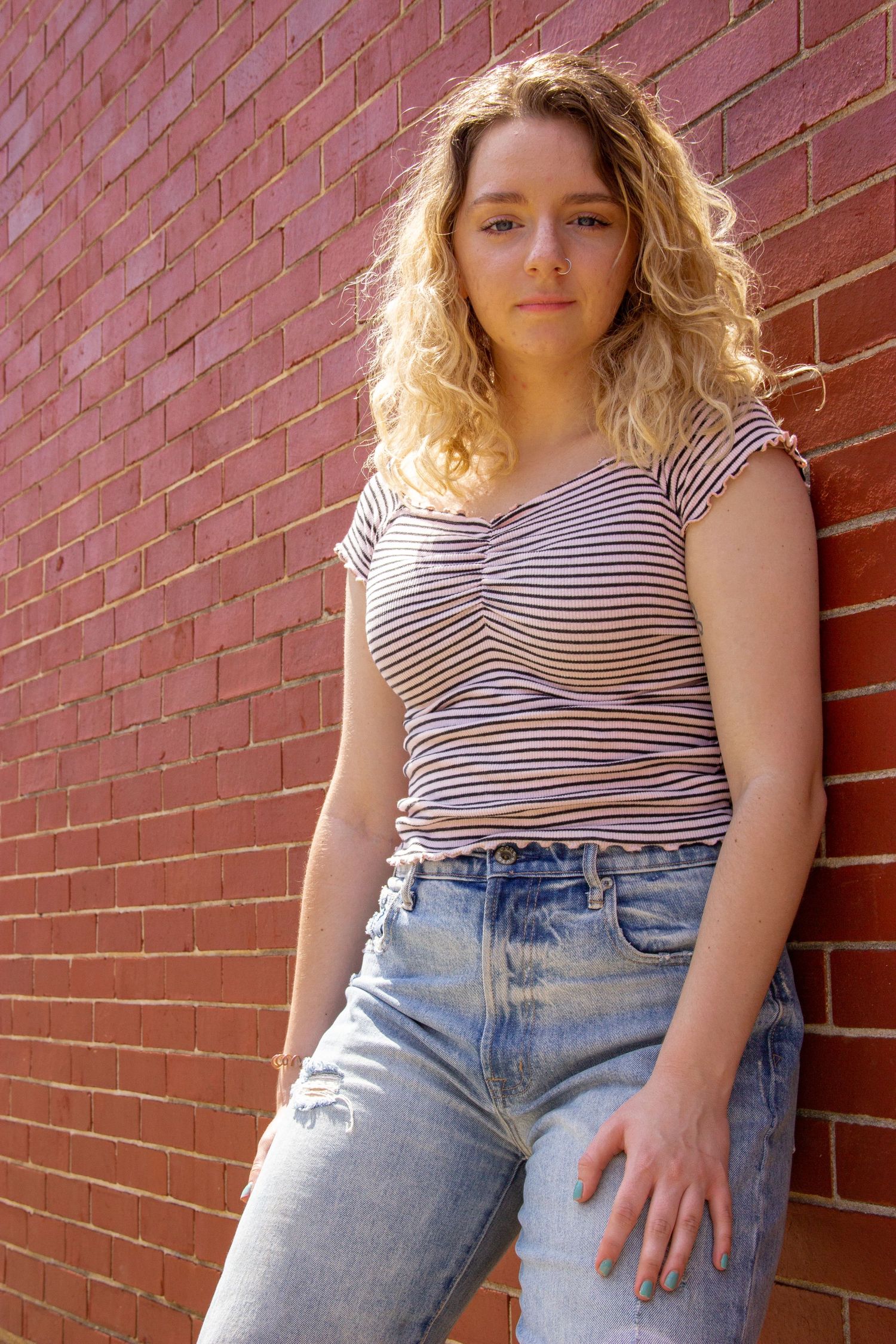 Young woman with curly blonde hair in striped top and ripped jeans against red brick wall.
