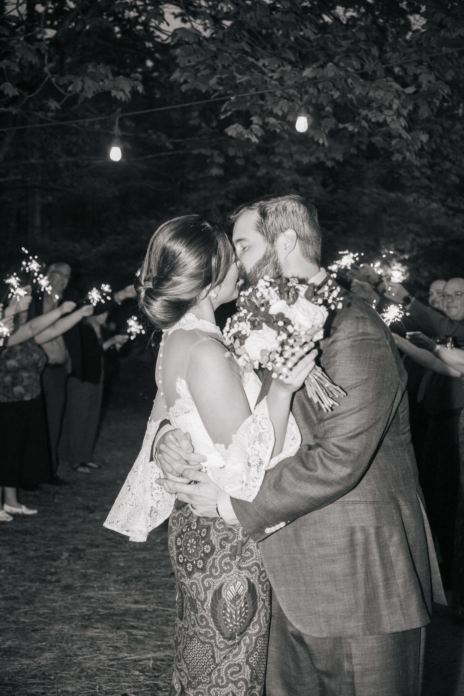 Black and white photo of newlyweds kissing during sparkler send-off at outdoor evening wedding reception.