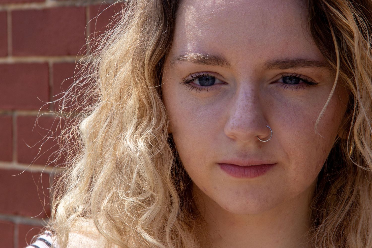 Close-up portrait of young woman with curly blonde hair and nose ring near brick wall.