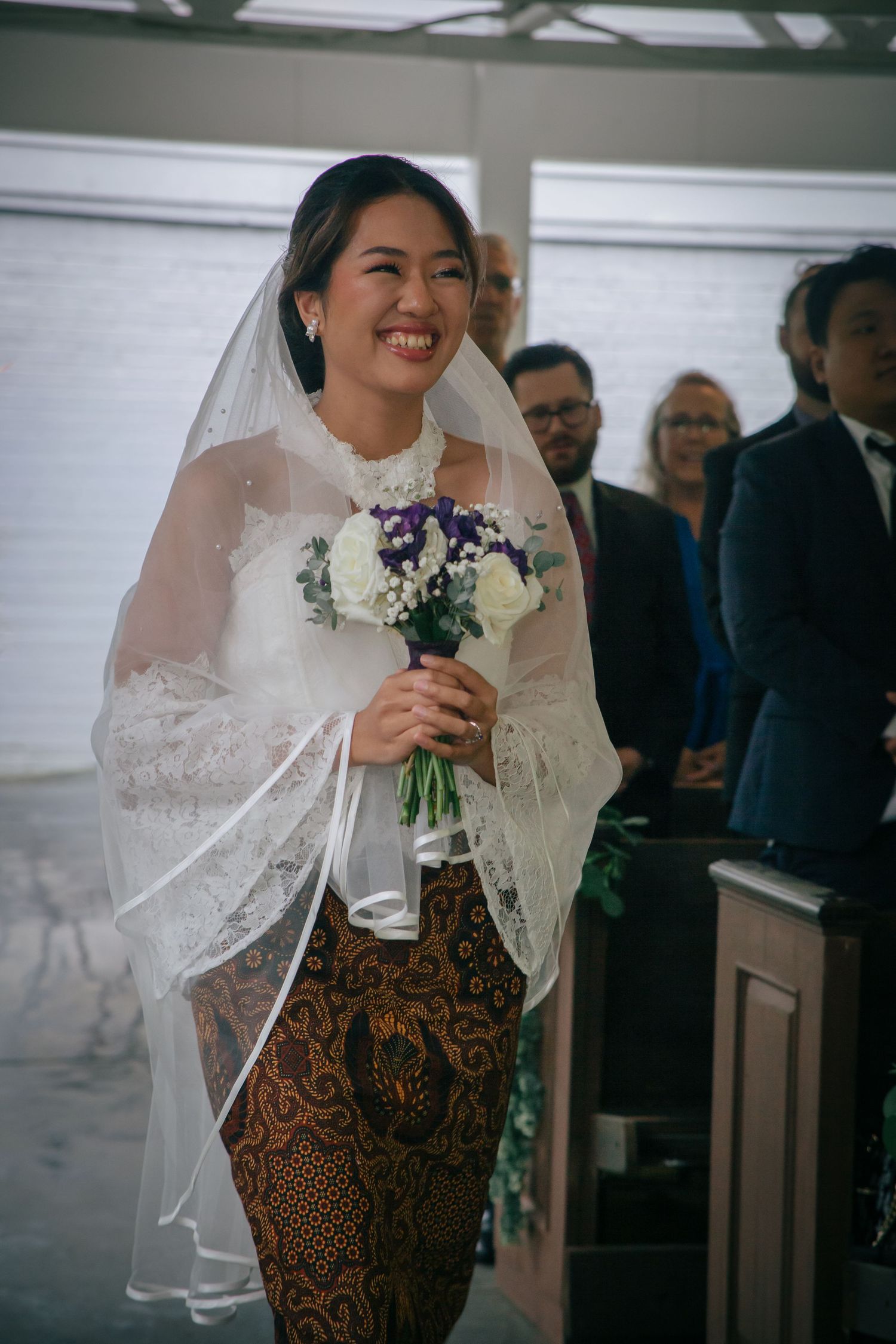 Radiant bride in lace kebaya and batik skirt walking down aisle holding purple and white bouquet.