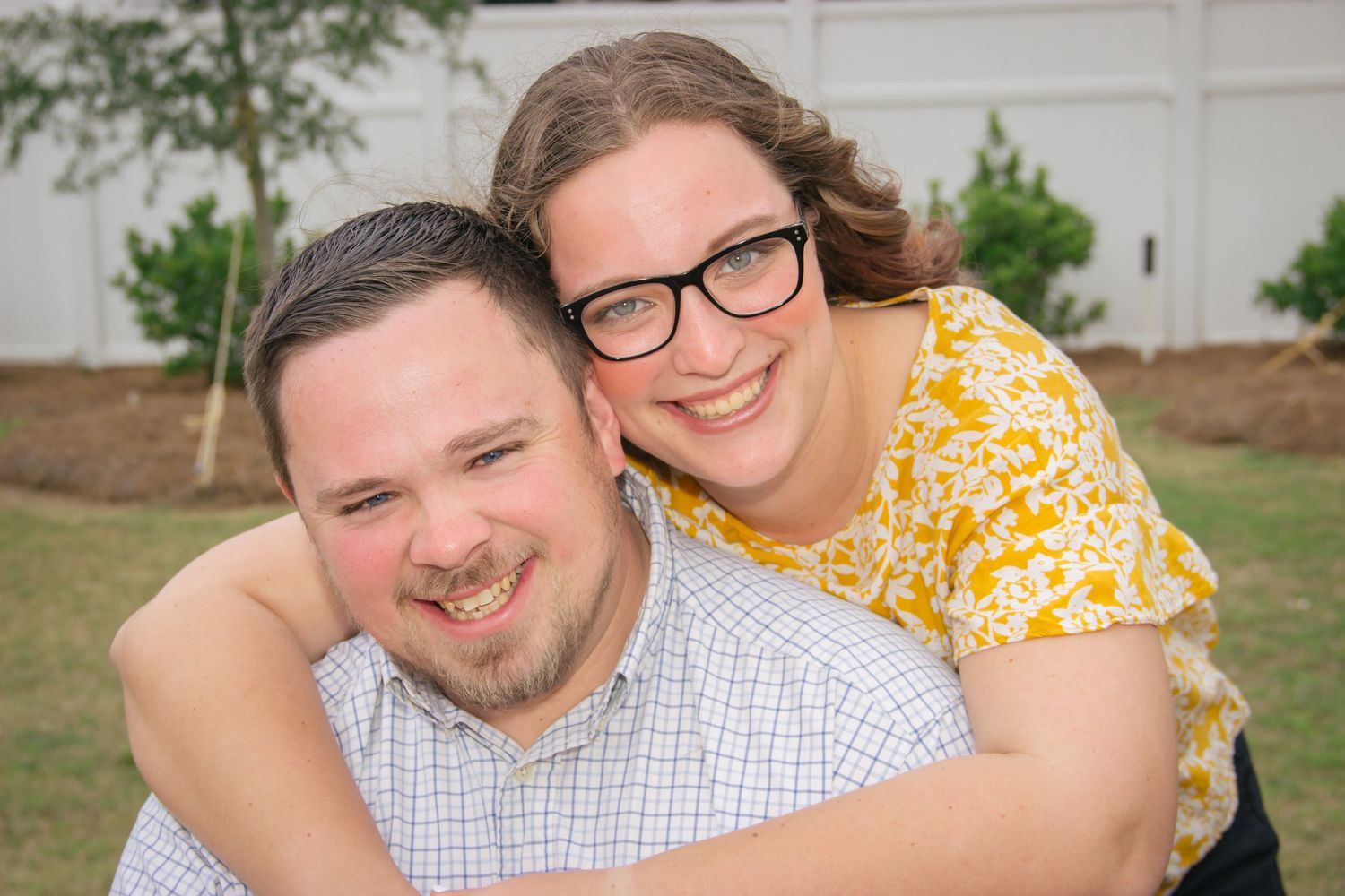 Happy couple embracing outdoors, woman with glasses hugging man from behind near white fence.