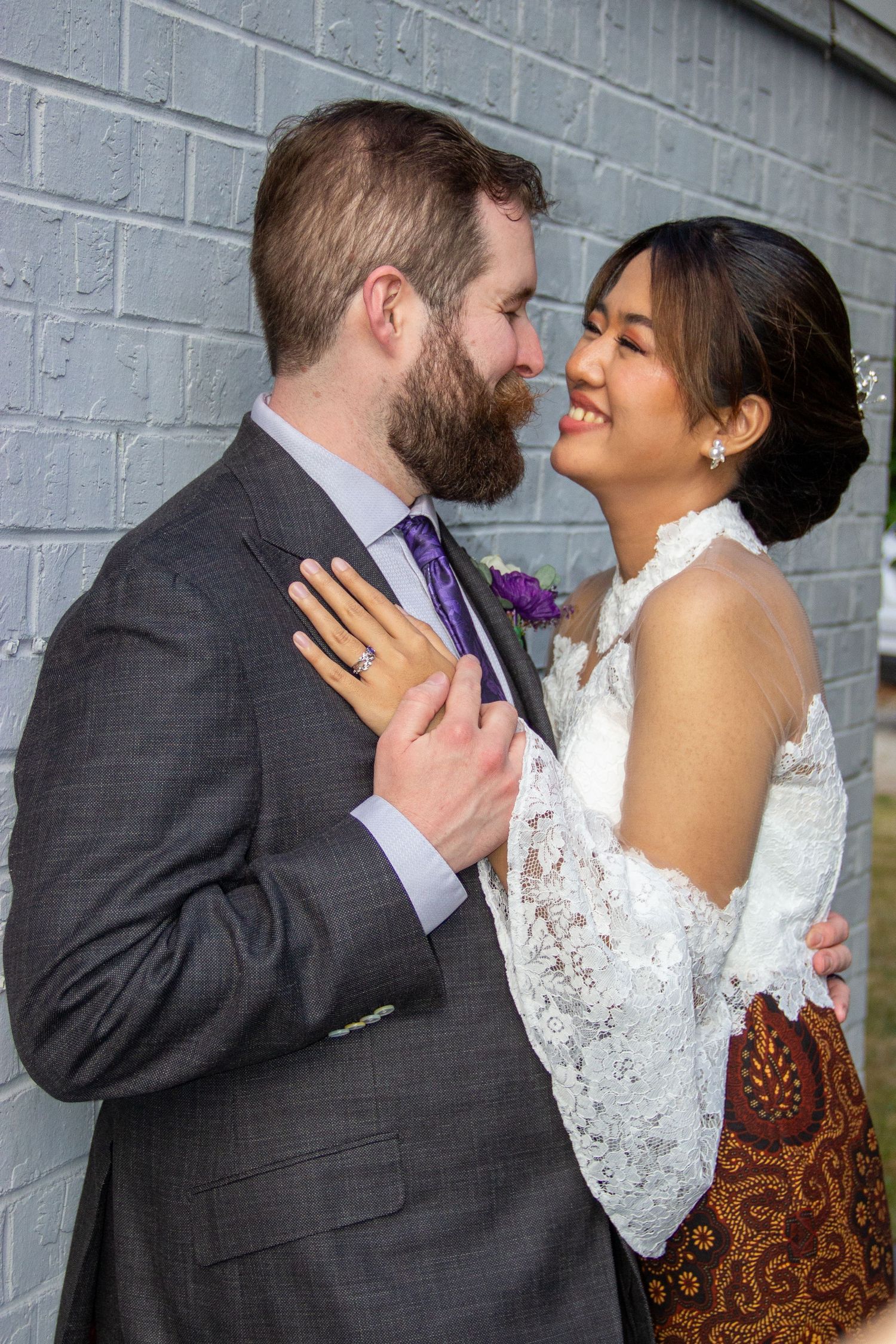 Bride in lace dress and groom in gray suit sharing romantic moment against gray brick wall.