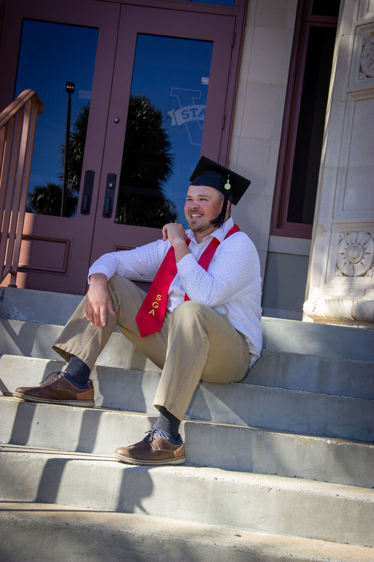 Graduate in cap and red stole sitting relaxed on stone steps outside campus building.
