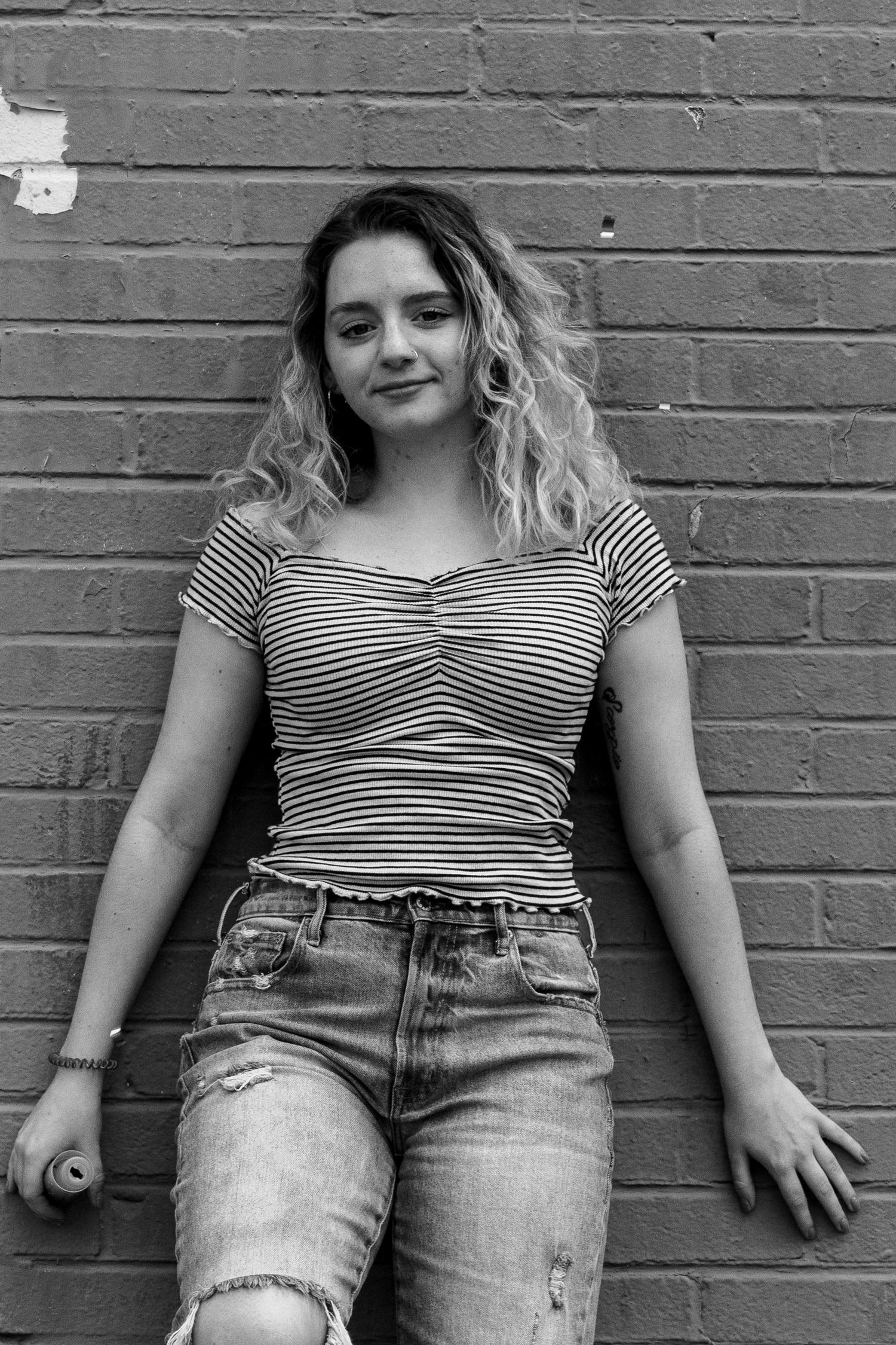 Young woman with curly hair in striped top and ripped jeans leaning on brick wall.