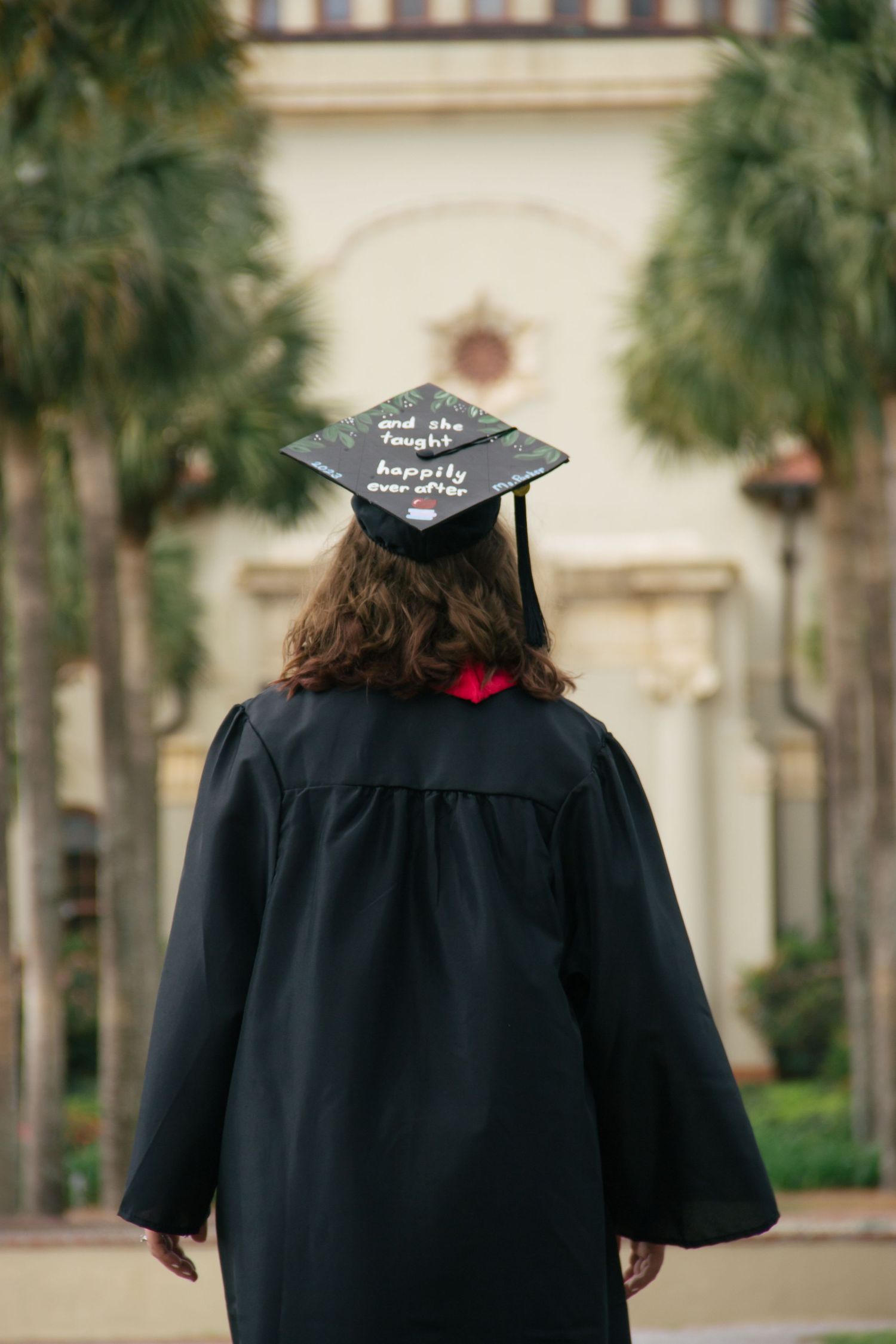 Back view of graduate in black cap reading and she lived happily ever after on campus path.