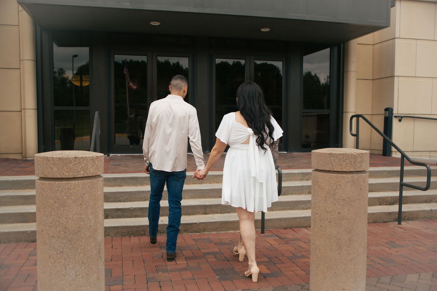 Couple holding hands walking toward modern building entrance during sunset engagement photo session.