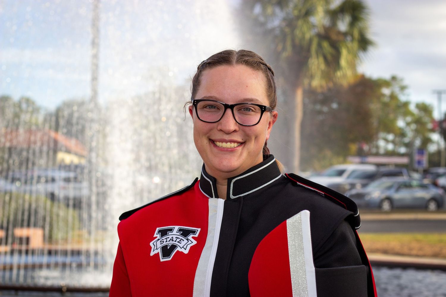 Smiling female marching band member wearing red and black uniform and glasses near fountain.