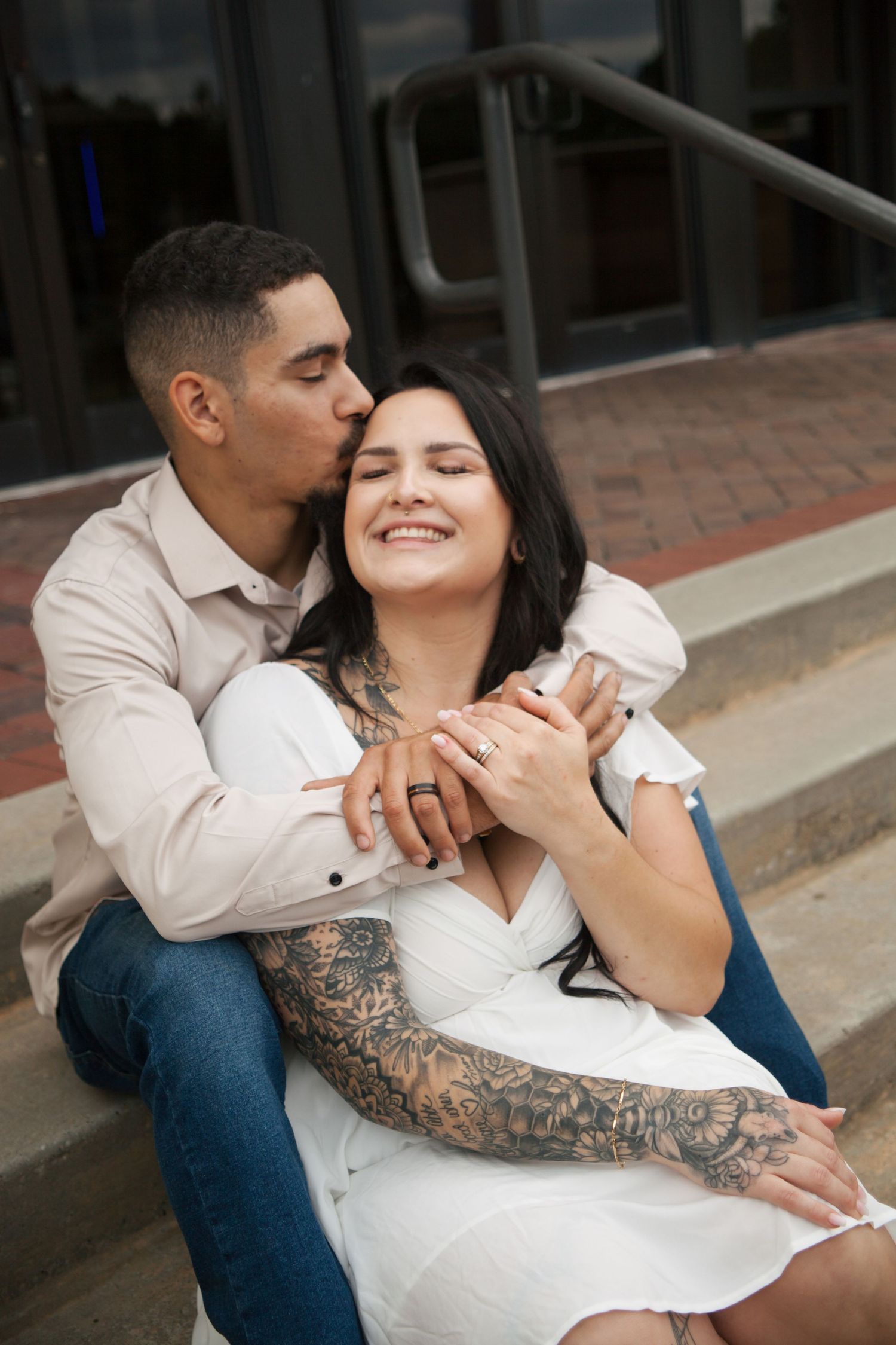 Couple sitting on steps, man kissing tattooed woman's cheek while she smiles with eyes closed.