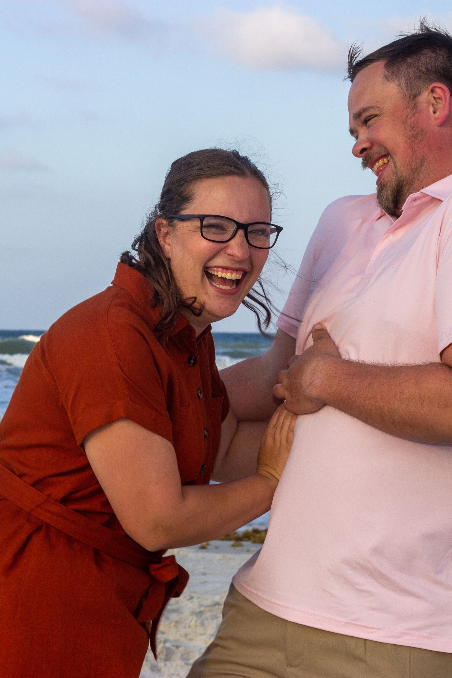 Laughing couple at beach, woman in red dress and glasses playfully grabbing mans side.