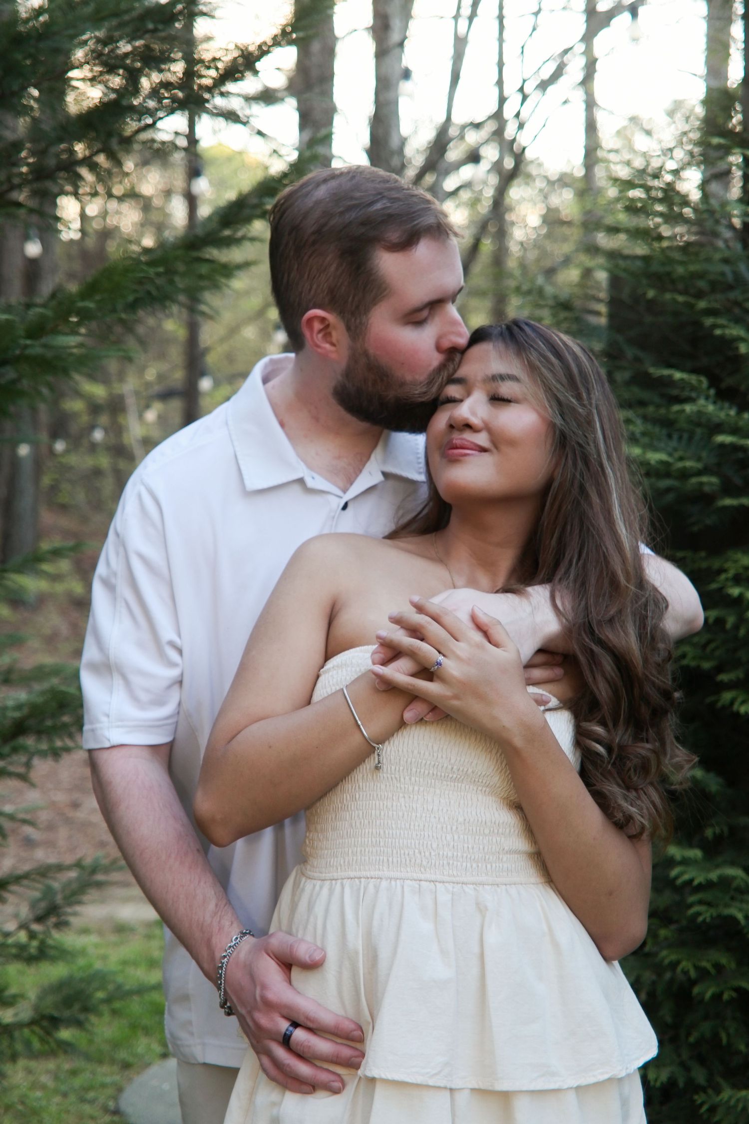Couple embracing in pine forest, man kissing woman's temple as she smiles in cream strapless dress.