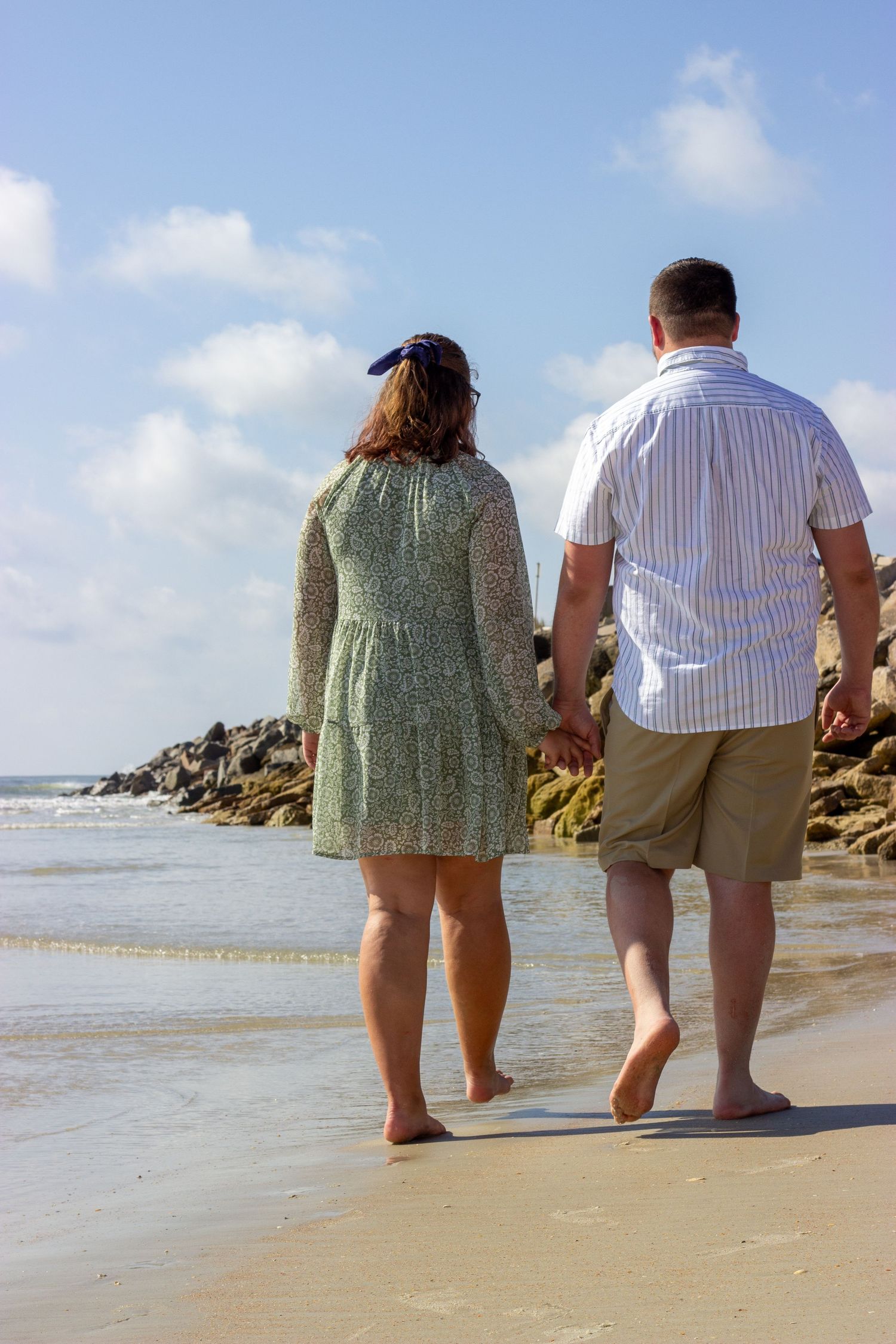 Couple holds hands walking barefoot along sandy beach on a sunny day.