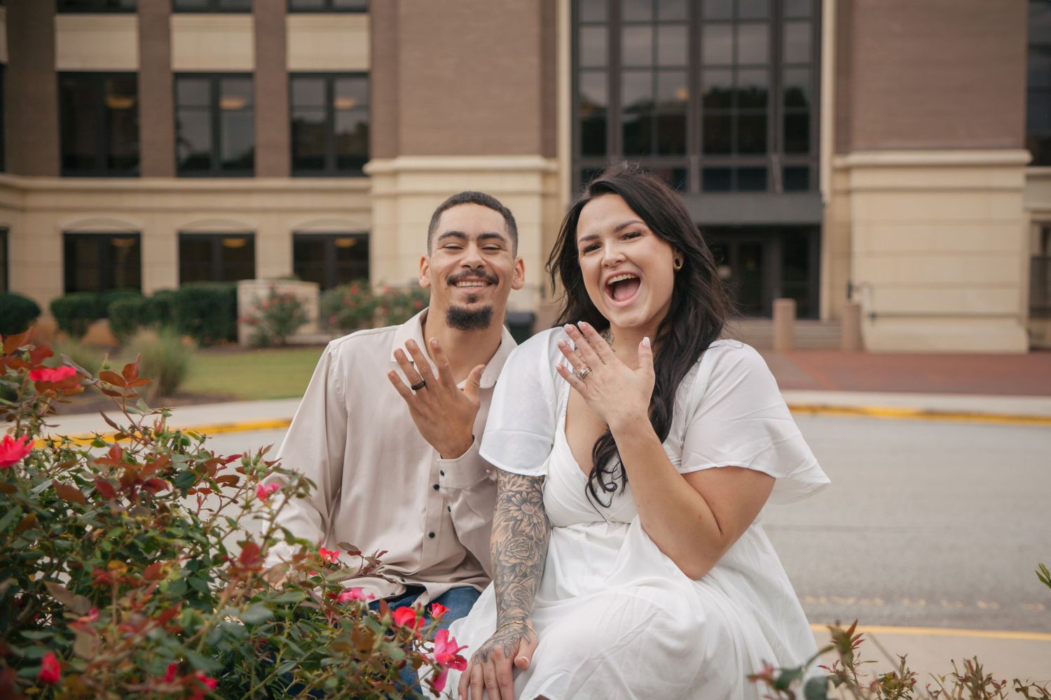 Excited engaged couple showing off rings while sitting among red roses outside urban building.