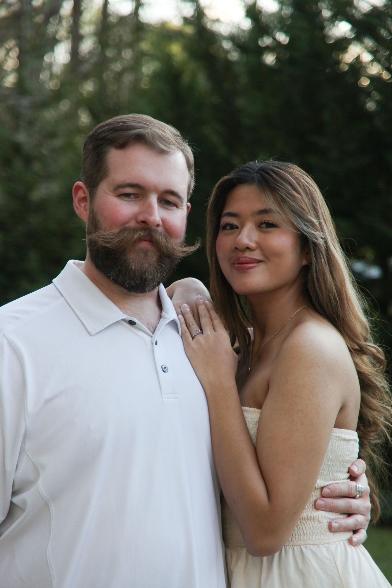 Bearded man and long-haired woman pose together outdoors in matching cream outfits during engagement photo session.