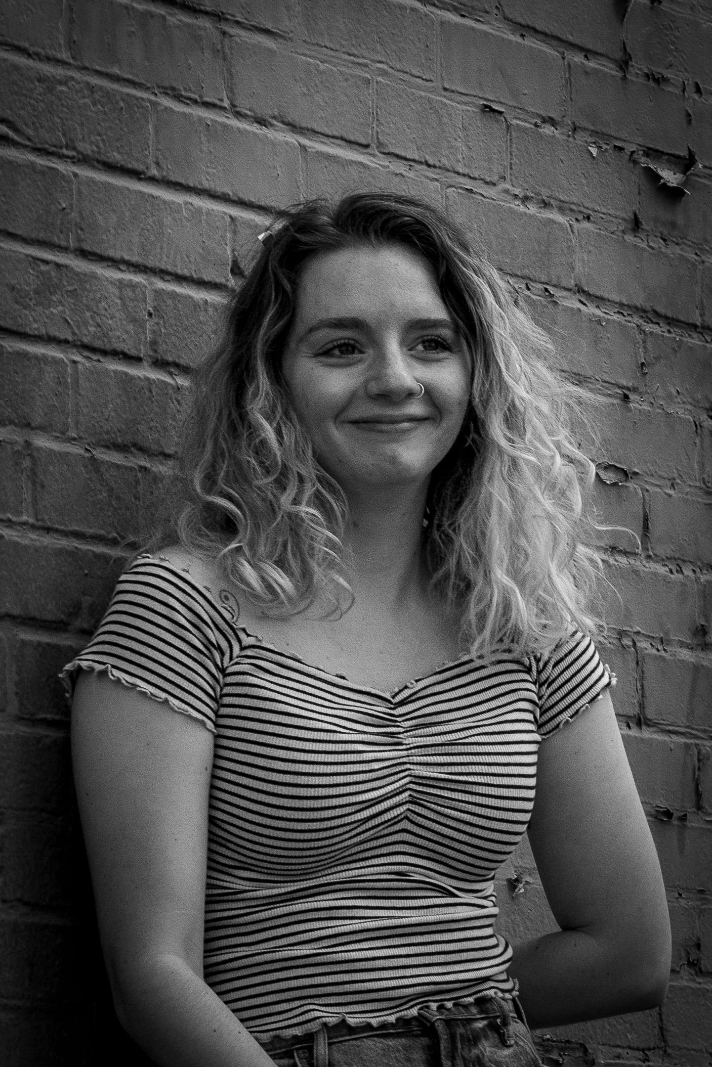 Black and white portrait of curly-haired young woman leaning against brick wall.