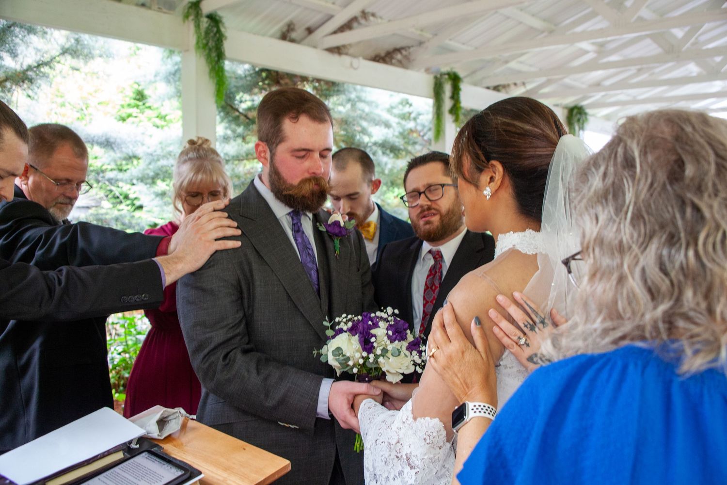 Wedding ceremony blessing with officiant and guests laying hands on bearded groom and veiled bride at outdoor altar.