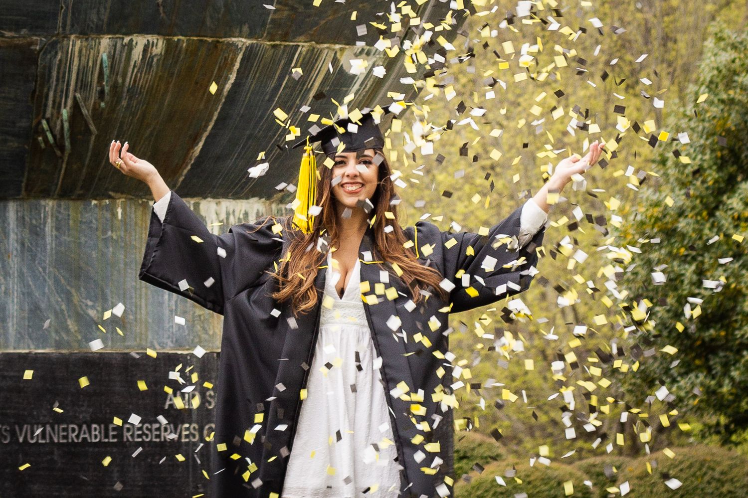 Joyful female graduate celebrating with gold and silver confetti in black graduation gown.