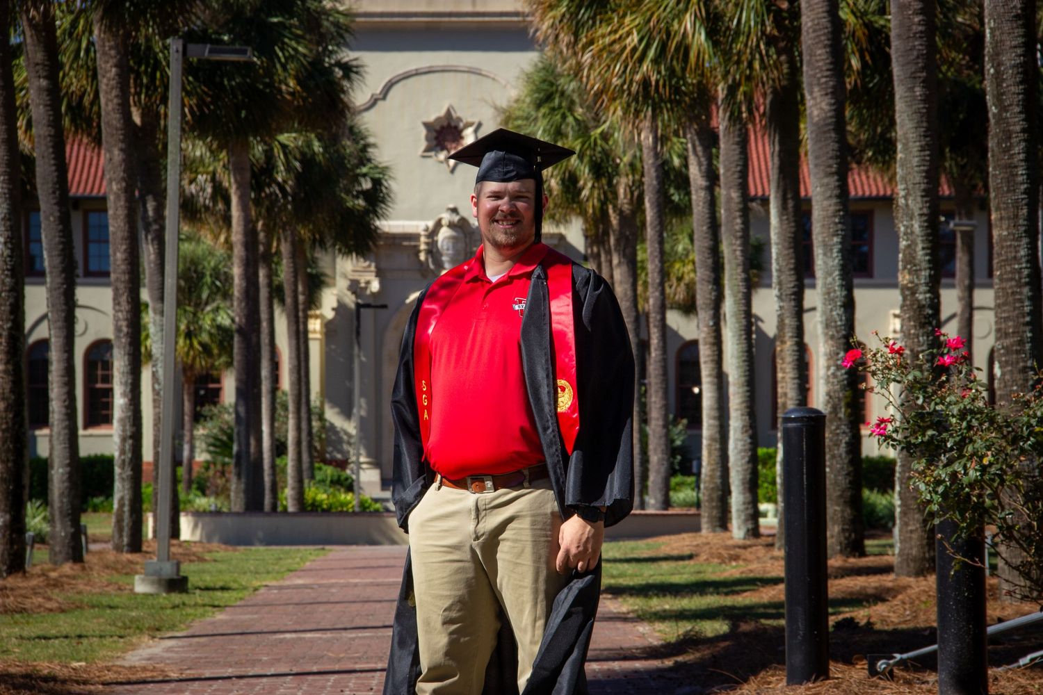 Male graduate in cap and gown walking along palm tree-lined campus pathway.