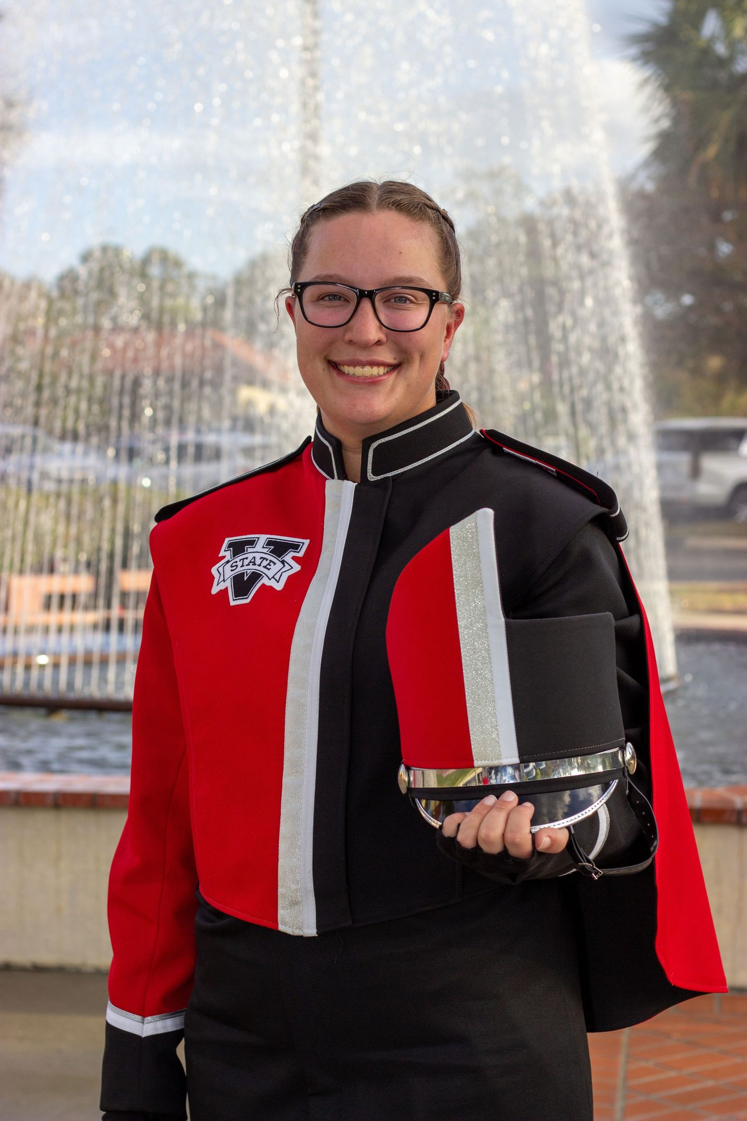 Smiling female marching band member in red black uniform holding hat near fountain.