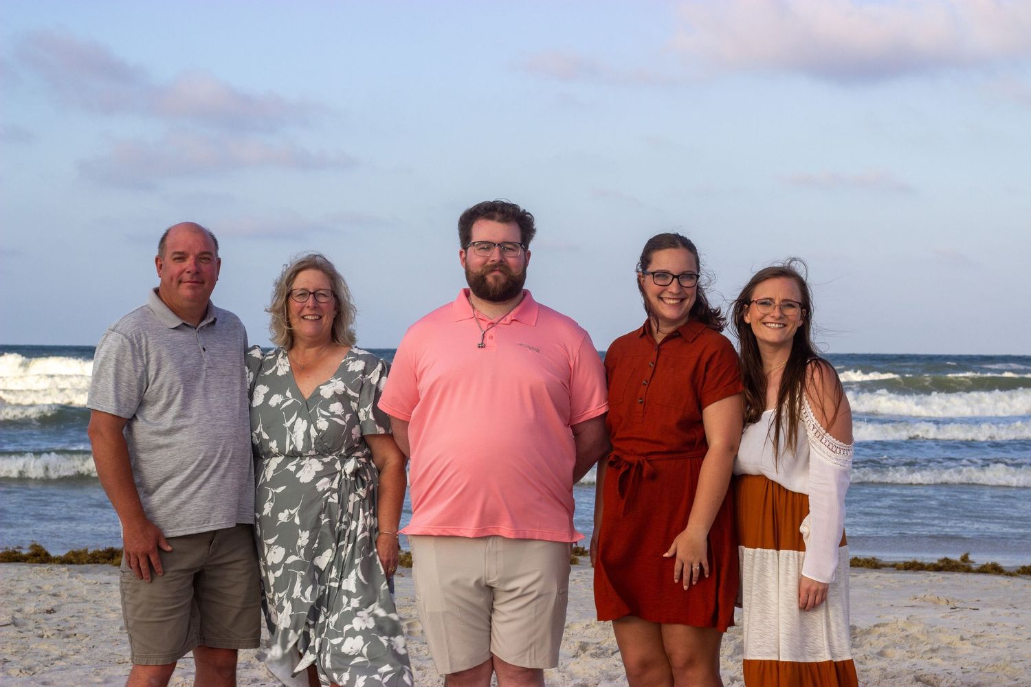 Family of five smiling together on sandy beach at sunset near ocean waves.
