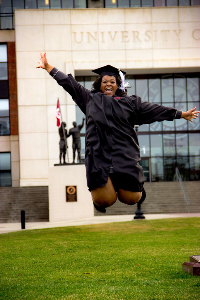 Excited graduate jumps joyfully in cap and gown outside university building.
