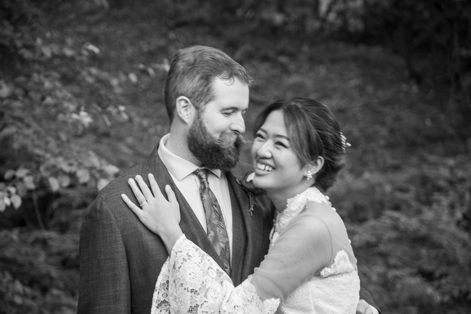 Black and white portrait of smiling bride in lace dress embracing bearded groom outdoors.