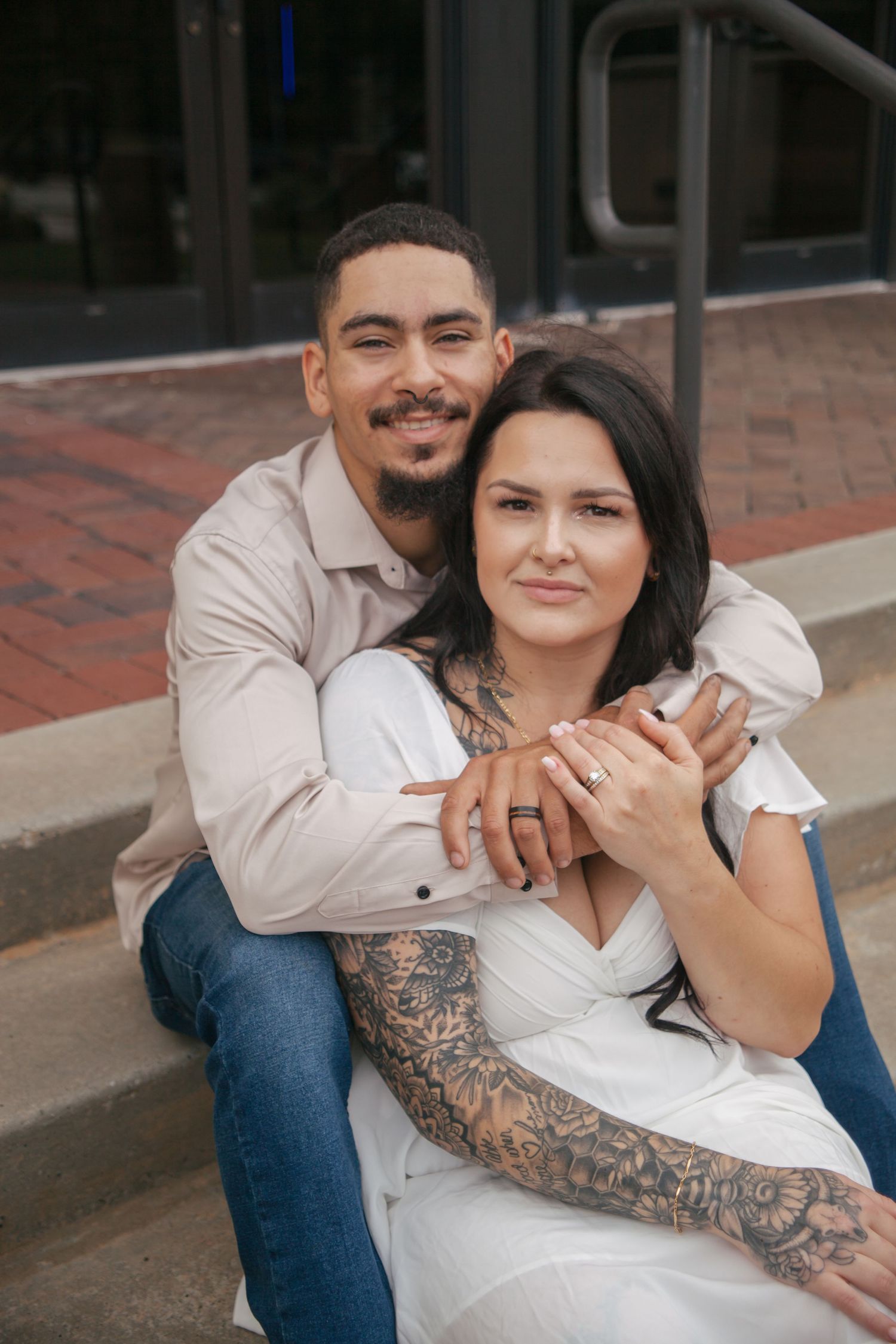 Tattooed woman in white dress and man in beige shirt pose together on brick steps during engagement session.