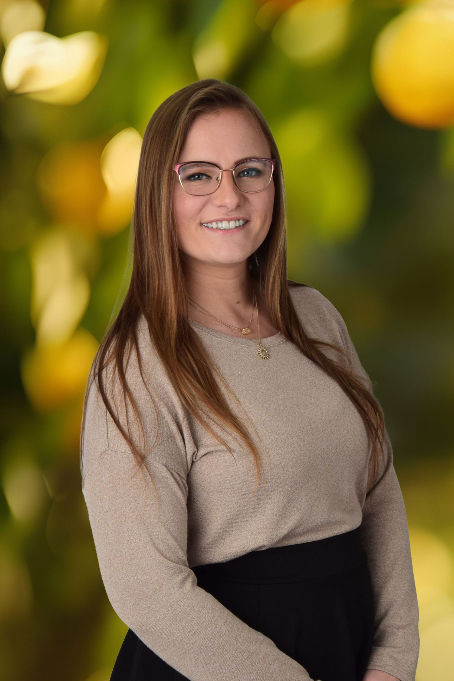 Smiling young woman with glasses poses outdoors with blurred yellow bokeh background.