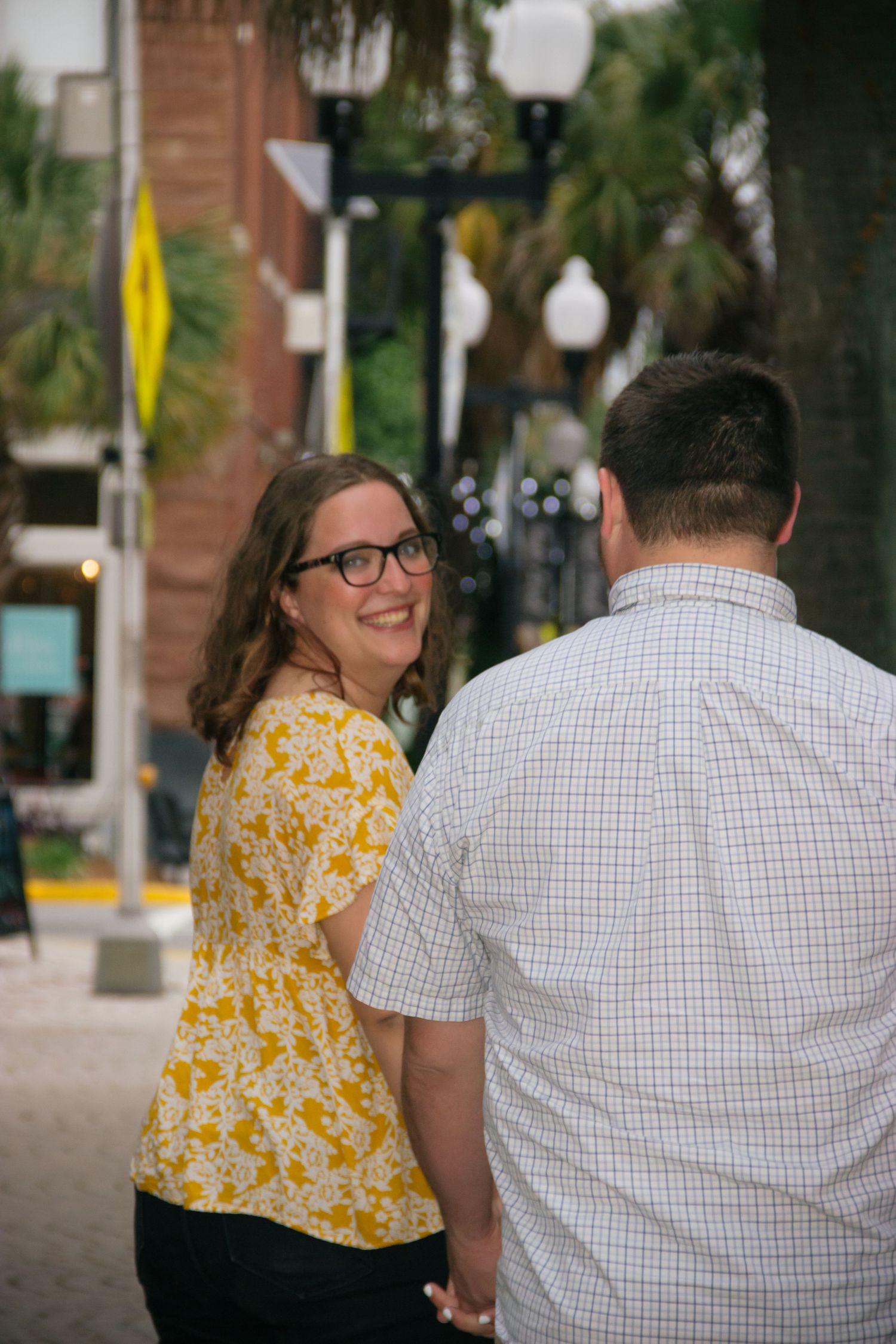 Woman in yellow floral top smiling back at camera while holding hands with man on urban street.