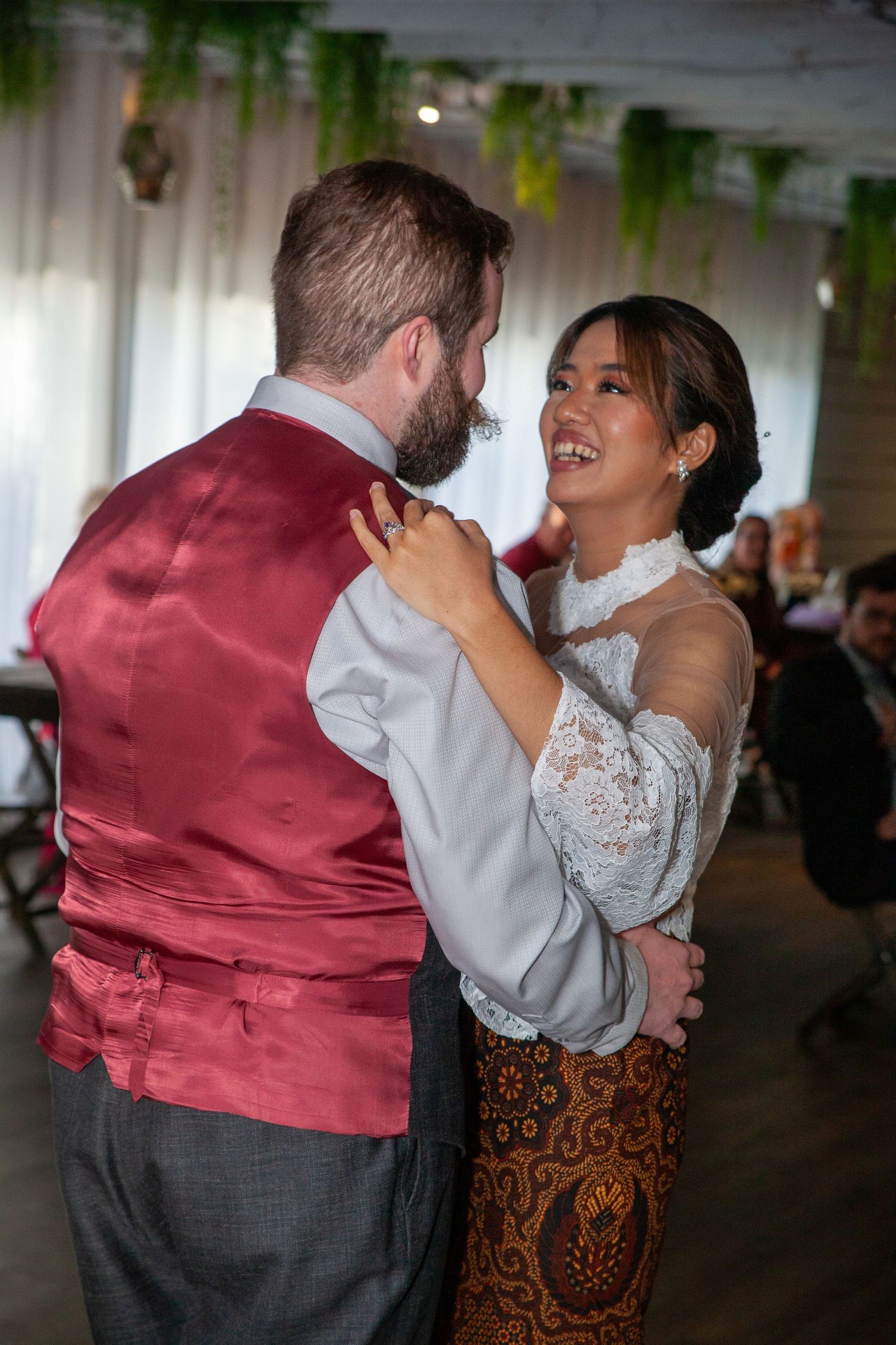 Smiling bride in lace top and batik skirt shares first dance with groom in red satin vest at wedding reception.
