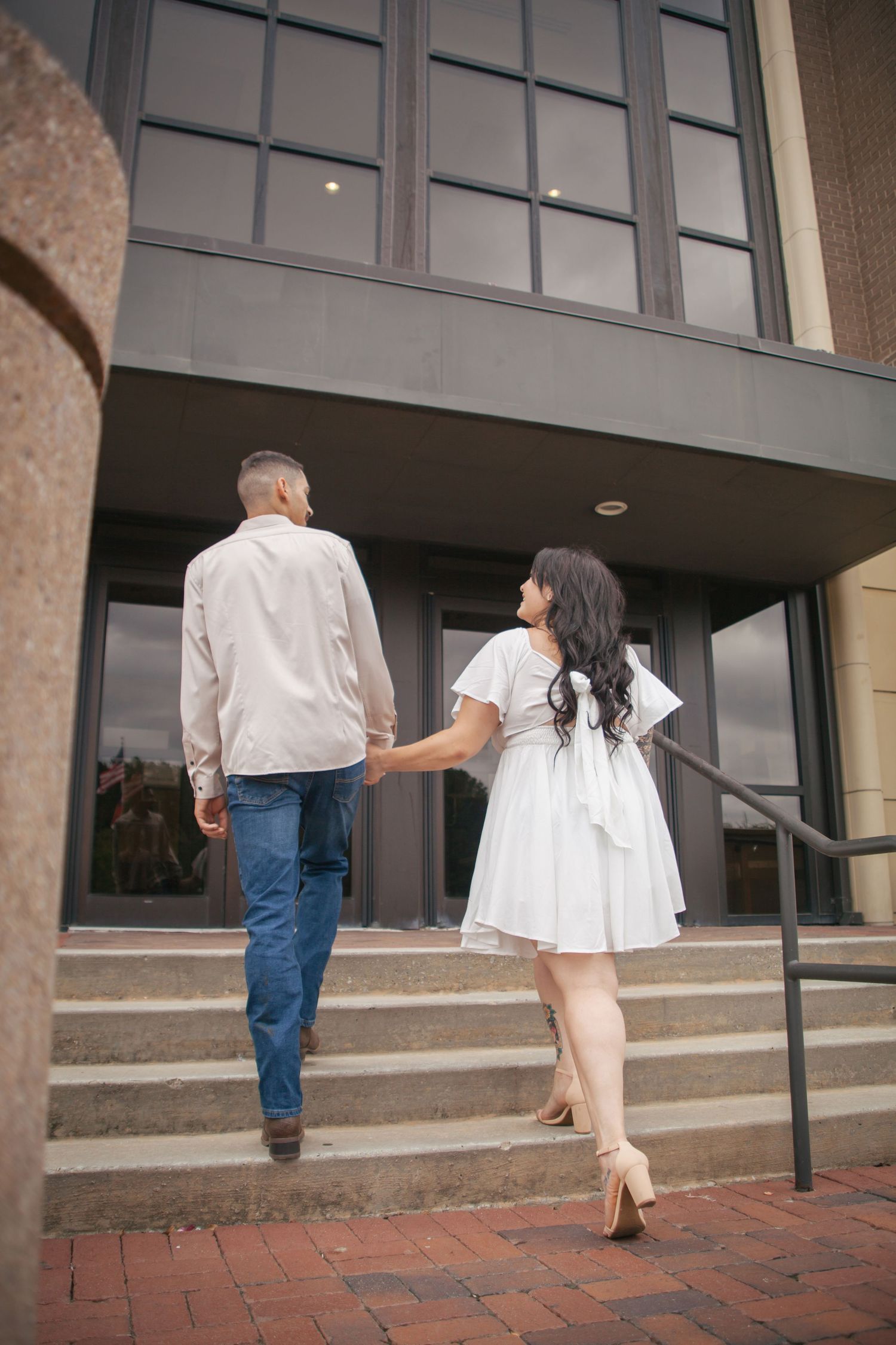 Couple holding hands walking up brick steps toward modern building entrance from behind.
