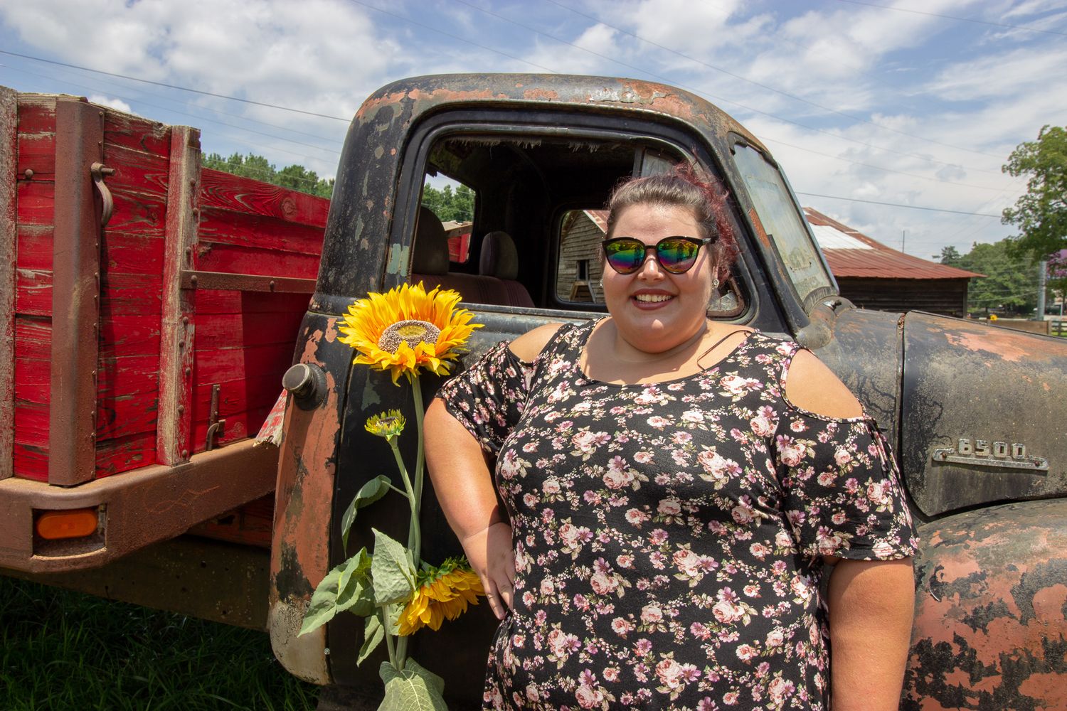 Woman holding sunflower poses beside vintage rustic truck outdoors on sunny day.