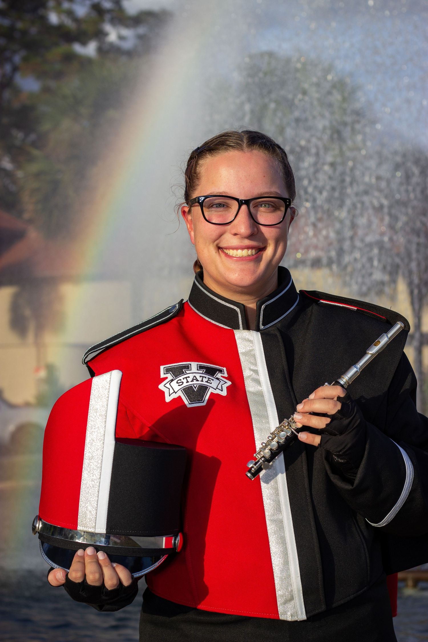 Smiling female marching band member holding flute and hat with rainbow behind her.