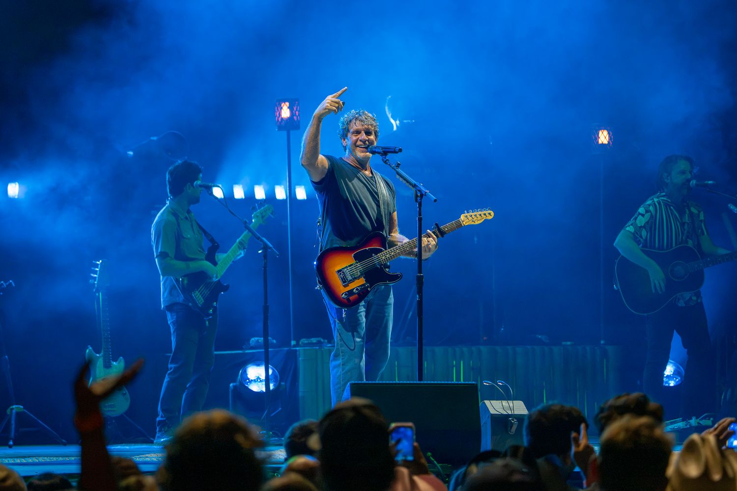 Rock band performing live on stage with blue lighting, lead guitarist raising hand to crowd during energetic concert.