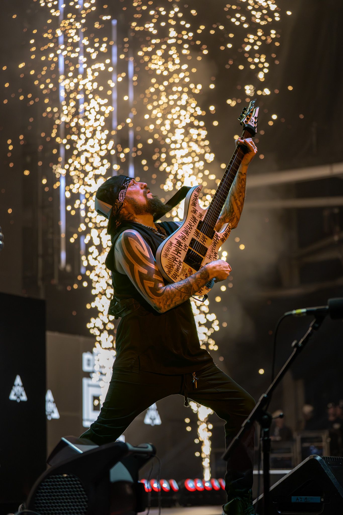 Tattooed guitarist performs on stage with golden confetti falling, raising electric guitar dramatically under concert lights.