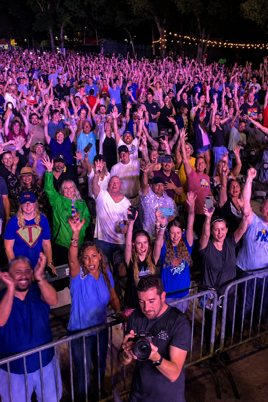 Excited crowd at outdoor concert at night, hands raised, photographer in foreground, string lights above.