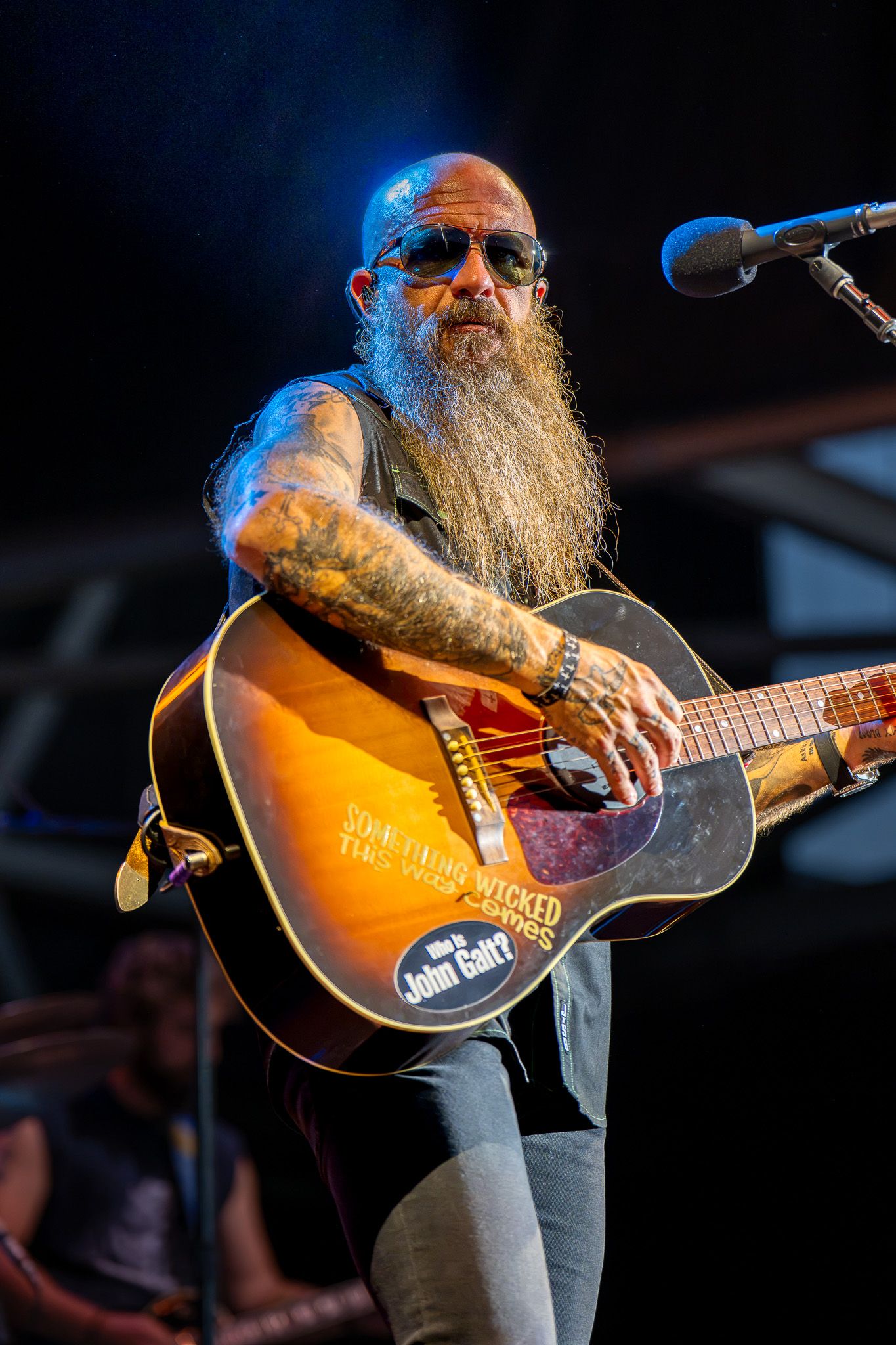 Tattooed musician with long beard playing acoustic guitar on stage near microphone during live outdoor performance.