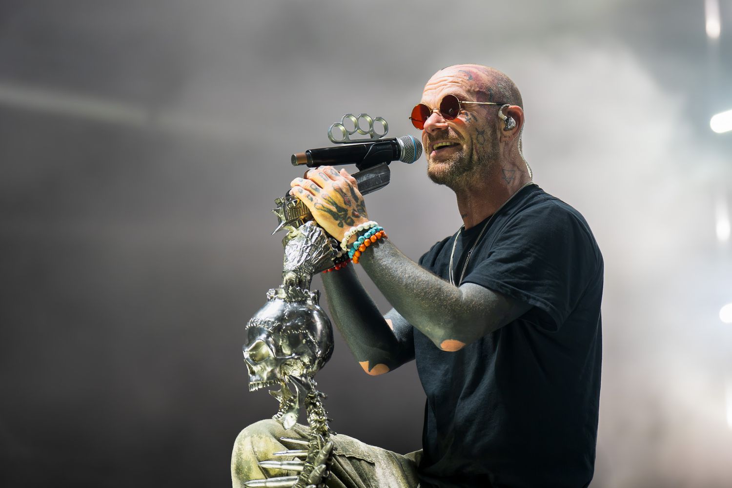 Tattooed male singer performing on stage, holding microphone near robotic sculpture, wearing black t-shirt and colorful bracelets.