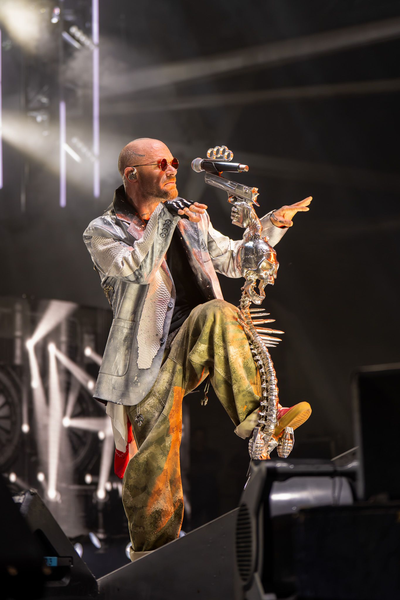 Bald performer in colorful tie-dye pants and silver jacket sings on stage holding microphone under dramatic lights.